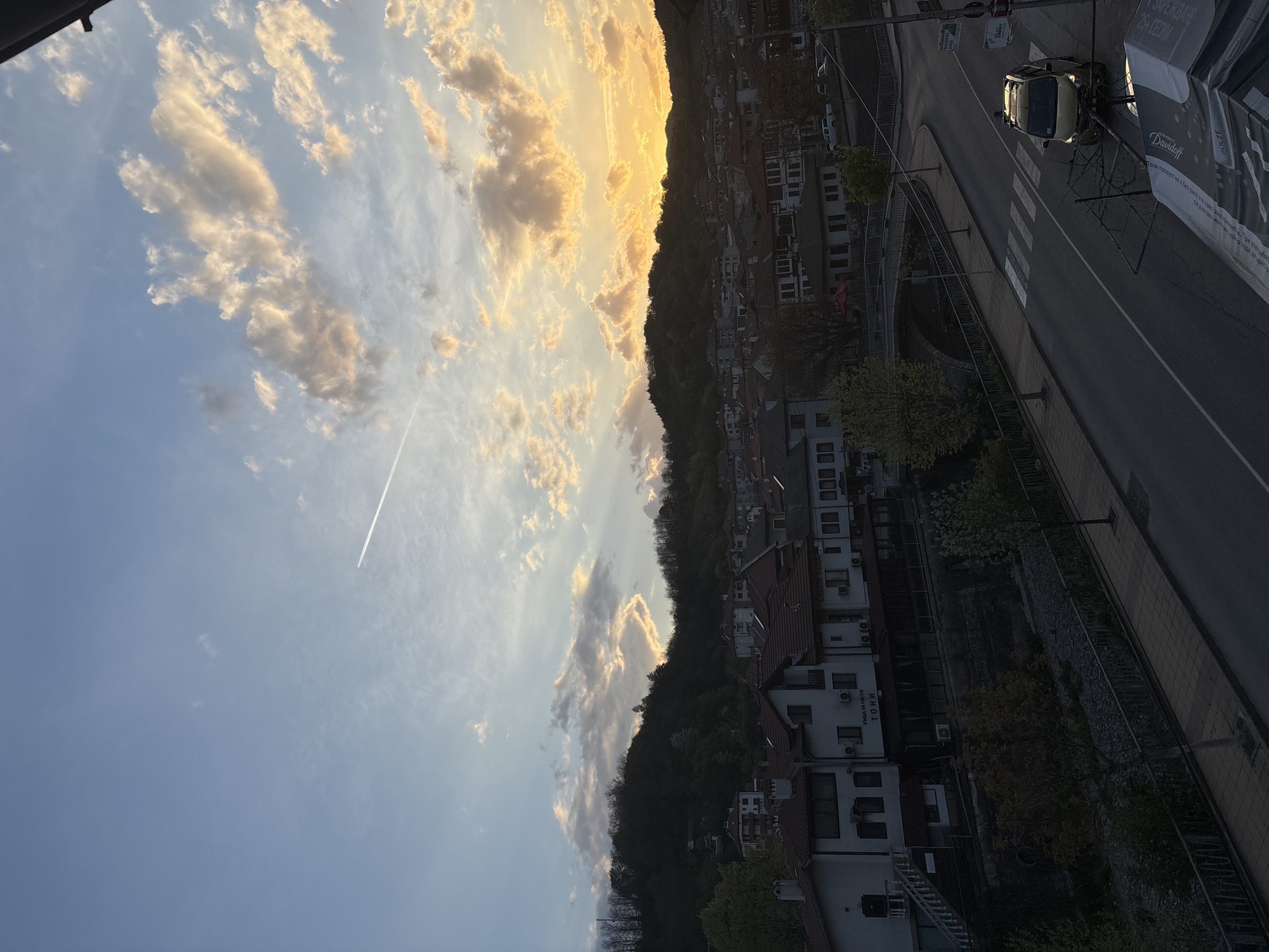 This image captures an aerial or high-angle view of the town of Zlatograd, Bulgaria, at either sunrise or sunset, bathed in a warm, golden light.
The upper portion of the frame features a dynamic sky. On the left, it transitions from a clear, pale blue to a more vibrant golden and orange hue towards the right, where the sun is low on the horizon. Scattered cumulus clouds, illuminated by the sun, display a range of light and dark tones. A prominent white contrail from an aircraft stretches across the blue part of the sky.
A dark, heavily forested hill forms a significant part of the landscape, silhouetted against the brightly lit sky. At the base of this hill, the town's architecture becomes visible, characterized by buildings with tiled roofs and mostly white or light-colored facades. Several buildings show external air conditioning units. A notable structure is a "КЪЩА ЗА ГОСТИ ТОНИ" (GUEST HOUSE TONI).
In the foreground and mid-ground, a paved road winds through the town, complete with a visible pedestrian crossing (zebra crossing). A light-colored car is parked on the side of the road. A bridge with a curved design is also discernible, suggesting it spans a river or ravine. Alongside the road, a large advertising banner is visible, displaying "PULZE" and "Davidoff," along with Cyrillic text including a health warning: "Този продукт не е без риск" (This product is not without risk), and promotional text like "ЗАРЕЖДАНЕ 25+ СЕСИИ" (CHARGING 25+ SESSIONS).
The overall impression is one of a tranquil town scene, likely at the start or end of the day, with no specific human activity evident, creating a peaceful ambiance.