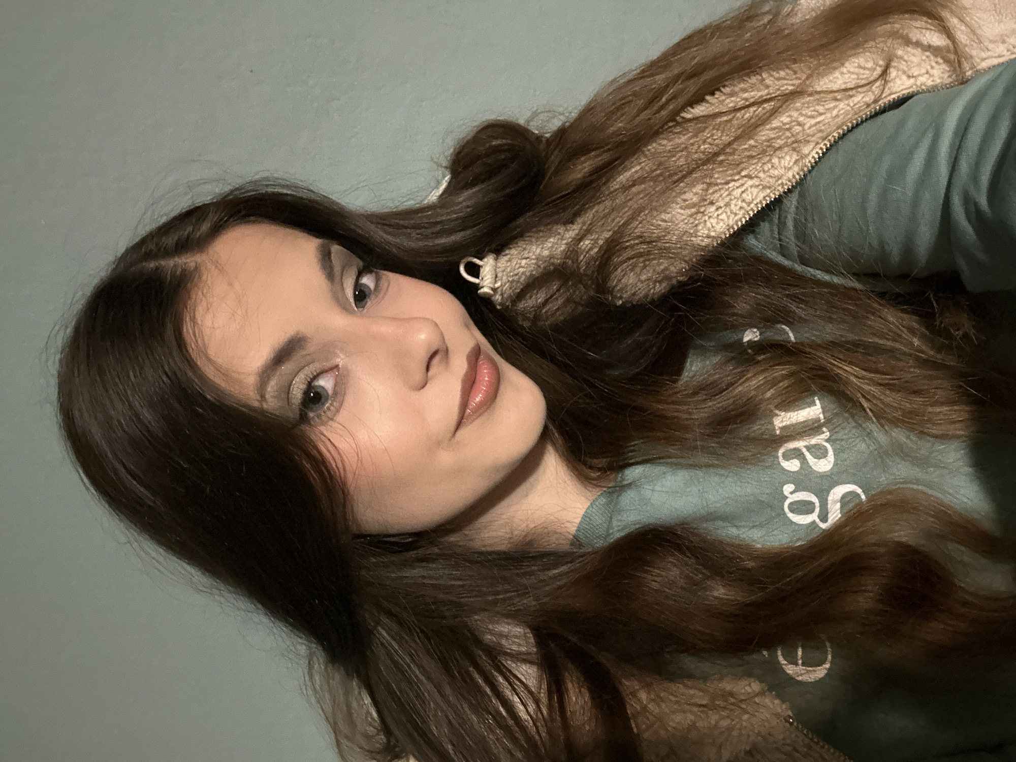 A young woman with long brown hair is the focus of this photograph. She is wearing a light green t-shirt with white lettering and a beige fleece jacket. Her makeup includes eyeshadow and lip gloss. The background is a solid, light blue color. The photograph is taken from a slightly low angle, and the woman is looking directly at the camera. The location is unknown.
