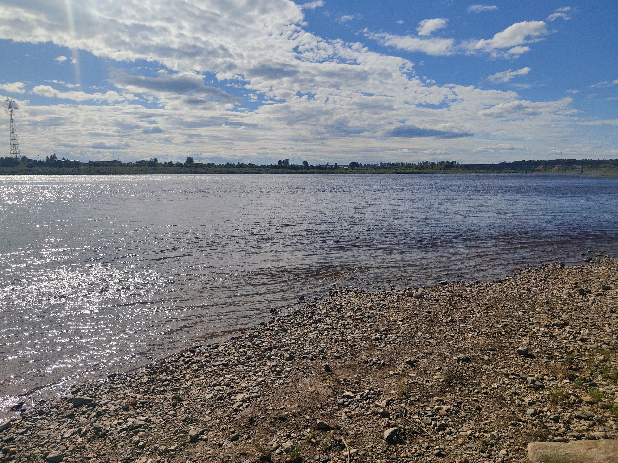 This media file depicts a scenic river landscape under a bright, partly cloudy sky. The foreground is a rocky shore with small to medium-sized stones, giving way to the calm surface of a wide river. Sunlight glints off the water, creating a shimmering effect. In the distance, the opposite bank is lined with a dense strip of green trees and vegetation. A tall, lattice-like power pylon is visible on the left side of the distant shore, and further to the right, a more subtle, possibly natural elevation or man-made structure can be seen. The sky is a vibrant blue, interspersed with fluffy white clouds, some of which are catching the sunlight. The overall impression is one of a peaceful, natural setting. The time of day appears to be late morning or early afternoon, given the bright sunlight and the position of the shadows, or lack thereof. The weather is clear and sunny, with a few clouds adding visual interest to the sky. There is no discernible text in the image. The scene conveys a sense of tranquility and the vastness of nature.