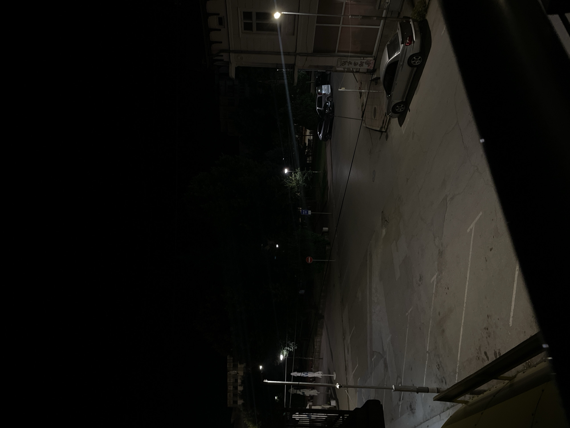 The image depicts a street scene at night in Pavlikeni, Bulgaria. A parking lot is visible with several parked cars, including a silver sedan, a silver minivan, and a dark-colored car. Streetlights illuminate parts of the area, casting shadows. On the right side of the image, a building with a light-colored facade and windows is partially visible, with some signage that is difficult to read. In the background, trees and other buildings are dimly lit by the ambient light. A red circular sign with a white horizontal bar, likely a "do not enter" or "no entry" sign, is positioned on a pole in the middle ground. The overall atmosphere is quiet and still, characteristic of a small town at night.