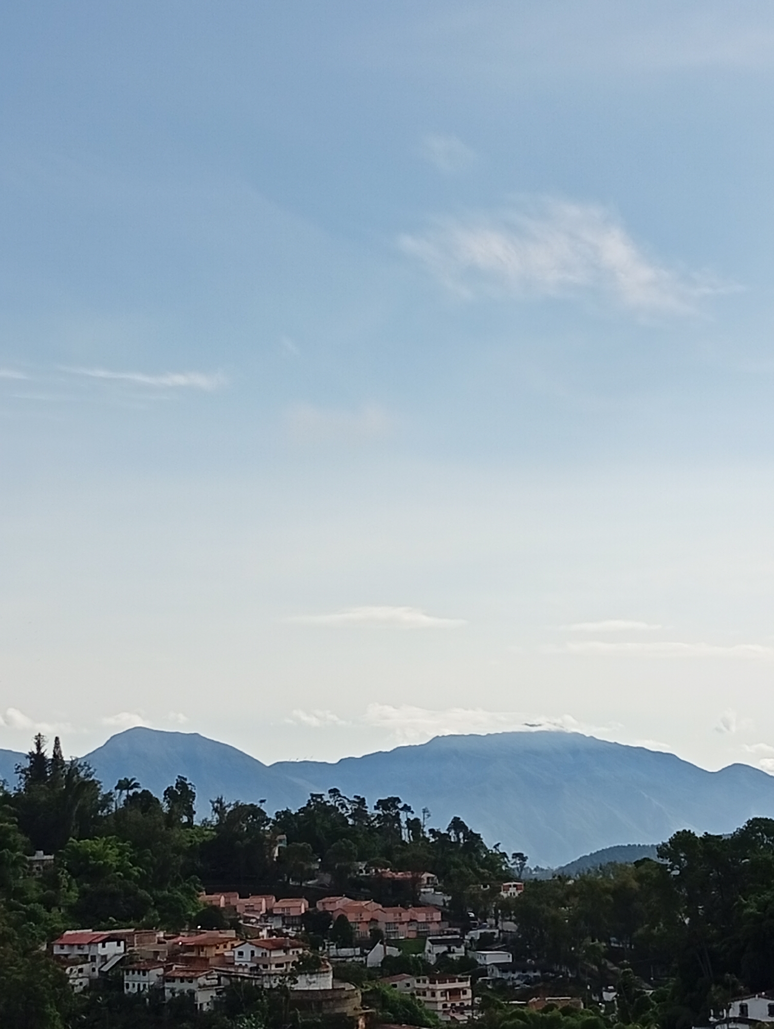 This is a landscape photograph taken from a high vantage point overlooking a residential area nestled against a backdrop of mountains. The sky is a pale blue with scattered wisps of white clouds, some of which are elongated and thin, suggesting a light breeze. The upper portion of the image is dominated by this expansive sky.
Below the sky, a range of hazy blue mountains stretches across the horizon, their peaks softly defined by the atmospheric perspective. Several layers of mountains are visible, creating a sense of depth.
In the mid-ground, a densely populated residential area is situated on a hillside. The houses are a mix of styles and colors, with many featuring red-tiled roofs. They appear to be relatively small to medium in size, and are packed closely together, indicating a suburban or urban development on the slopes. Greenery, consisting of trees and bushes, is interspersed throughout the residential area and on the surrounding hillsides, providing a natural contrast to the man-made structures. Some of the trees are tall and slender, while others are more bushy and full.
The foreground is also characterized by lush green foliage, suggesting that the photograph was taken from a higher elevation, looking down upon the scene. The overall impression is one of a town or city that has grown organically on the challenging terrain, with nature and human habitation coexisting. The lighting suggests it is daytime, likely morning or afternoon, with soft, diffused light. There are no visible people or animals in the frame. The scene captures a view of San Antonio de Los Altos, Venezuela, known for its hilly terrain and scenic mountain views.