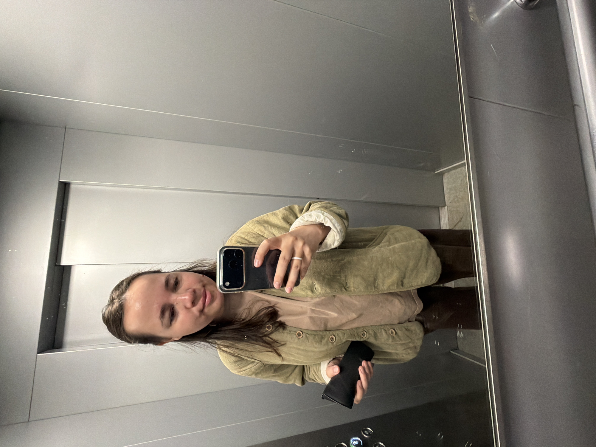 A woman is taking a selfie in a mirror inside an elevator. She is wearing a light green jacket and holding a black smartphone. The elevator has gray metal walls and control buttons visible near the bottom. The location cannot be determined from this image.
