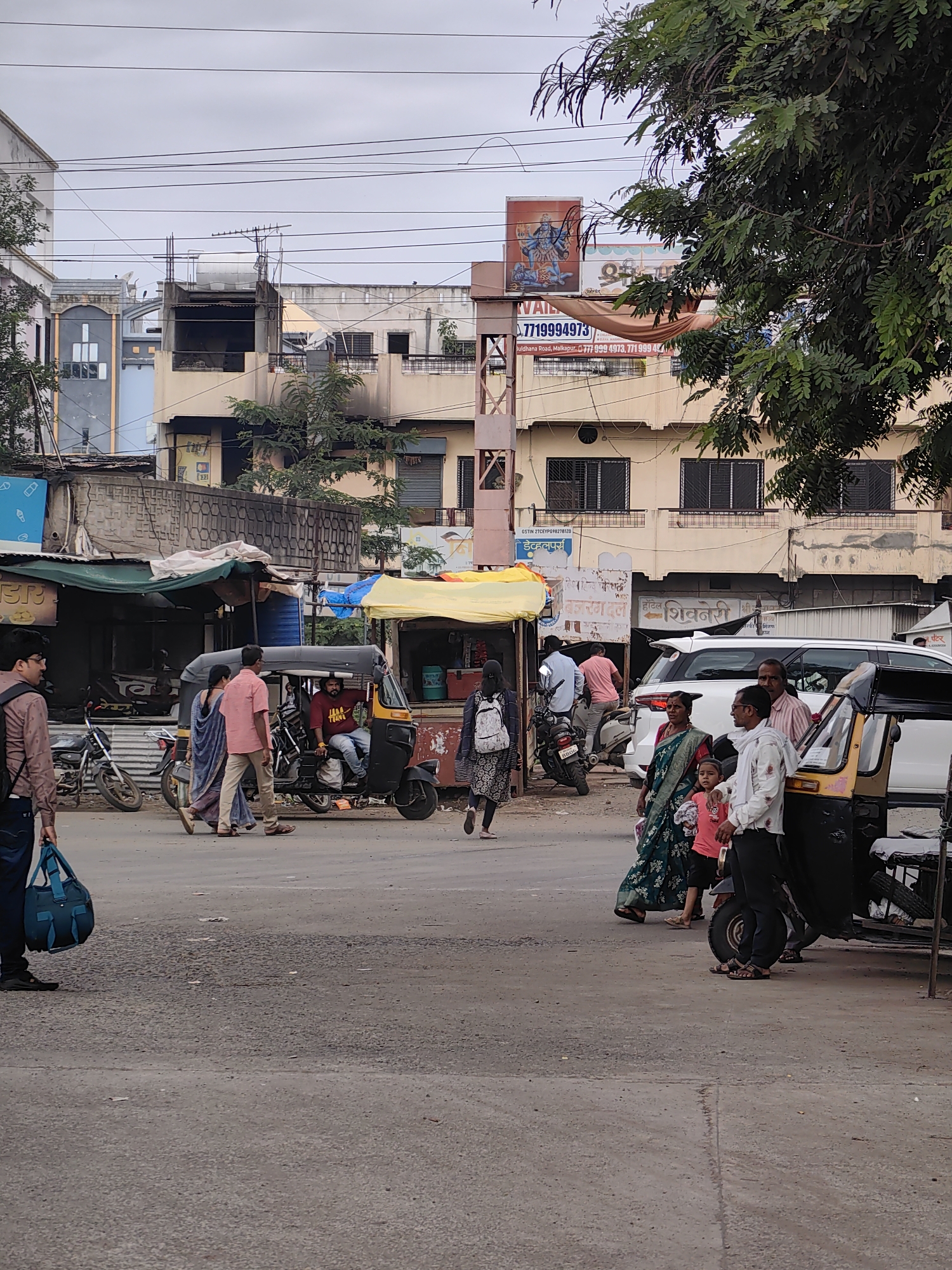 The image depicts a street scene in Malkapur, India, likely near a market or commercial area, with buildings lining the background and vehicles and pedestrians occupying the foreground. In the center of the frame, a yellow and black auto-rickshaw is parked, with a man wearing a red t-shirt driving it. Several people are visible around the auto-rickshaw and on the street. To the left, a man in a brown shirt and dark pants carries a blue duffel bag. Two women, one in a blue saree and the other in a pink outfit, walk past a parked motorcycle. Further to the right, a white SUV is parked, and a group of people, including a woman in a green saree and a child, stand near it, along with a man in a white shirt and dark pants. The street is paved with asphalt, and there are several shops and stalls with awnings. Overhead power lines crisscross the sky, which is overcast. A large tree with green foliage is present on the right side of the image, partially obscuring the buildings. The architecture of the buildings in the background appears to be a mix of traditional and more modern styles. A billboard with an image of a deity and text in an Indian script is visible on one of the buildings.