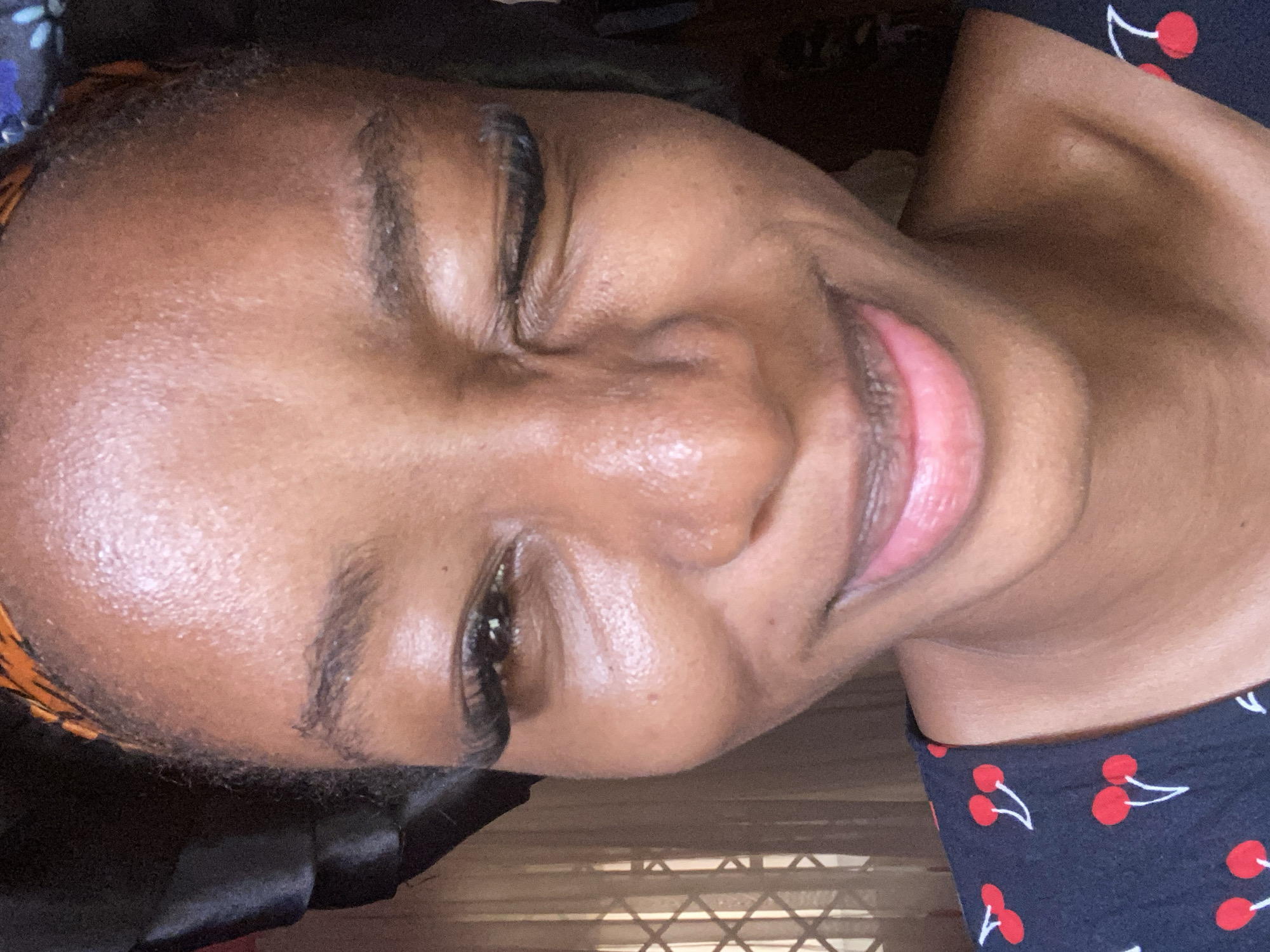 A close-up portrait in the city of Jos, Nigeria, features a young individual with medium-dark skin, looking directly into the camera. The subject is winking with their proper left eye (viewer's right eye closed) and smiling with closed lips. Both eyes are adorned with prominent false eyelashes. The eyebrows are naturally dark and full. The skin on the forehead, cheeks, and nose exhibits a noticeable sheen or gloss. The lips are a natural pinkish-brown tone. The individual wears a dark-colored garment, possibly navy blue or black, with a repeating pattern of white-stemmed red cherries. A head covering is visible, featuring an orange and black patterned fabric at the crown and a black satin-like material on the proper left side of the head. The background on the proper left (viewer's left) side reveals a window with a visible metal security grill or patterned translucent curtains, suggesting natural light is entering from this direction. The background on the proper right is darker and out of focus.