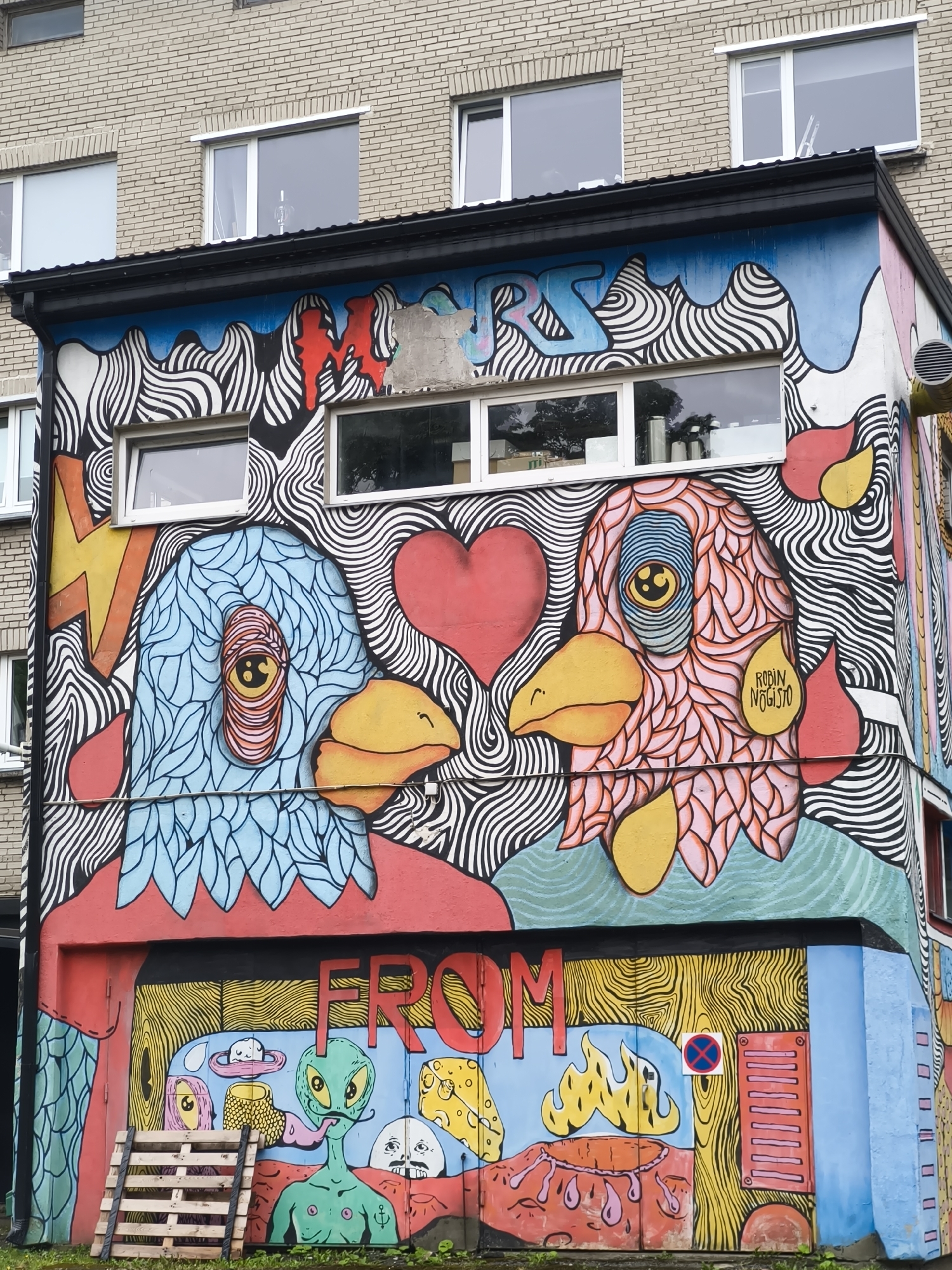 The image shows a colorful mural painted on the side of a building in Tallinn, Estonia. The mural features two stylized bird-like heads facing each other, one in blue and the other in pink, with large, expressive eyes. Both birds have yellow beaks and are surrounded by swirling black and white patterns. Above the birds, partially obscured by paint damage, are the letters "MR." Below the birds, the word "FROM" is painted in large red letters. The lower part of the mural depicts various whimsical elements, including a green alien with a long tongue, a stack of what appears to be cheese, and a pink hat with a white orb on top. A wooden pallet lies on the ground in front of this section. The building itself appears to be made of light-colored bricks and has several windows. The mural is vibrant and detailed, with a distinct street art style.