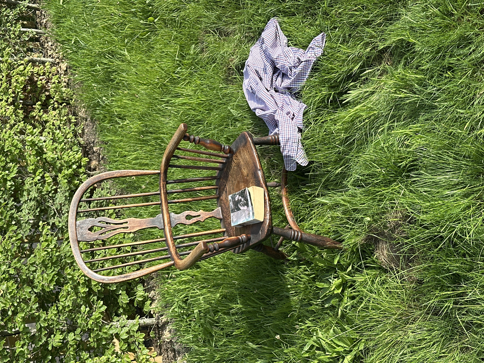 An eye-level shot depicts a wooden chair resting on grassy terrain outdoors. The chair is angled, its back facing the upper left corner of the frame. A book sits on the chair's seat, and a checkered shirt is draped over the chair's back and into the grass. The grass is green and lush, and an area of dense vegetation is visible in the lower left corner.