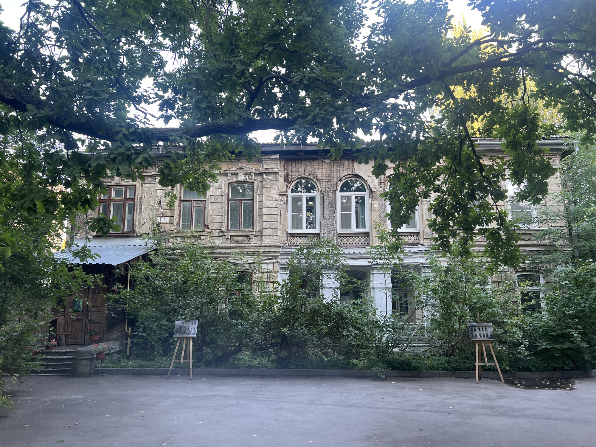 This is a medium shot, taken outdoors, of an old, weathered building in Nizhny Novgorod, Russia. The building, likely a residential or administrative structure, exhibits signs of decay, with crumbling plaster and exposed brickwork. It features several windows, some with arched tops, and a covered entrance on the left. The facade is adorned with decorative elements, suggesting a past architectural grandeur.
The building is partially obscured by lush green foliage, including a large tree with thick branches and dense leaves that extend across the top of the frame, casting shadows on the building. Bushes and smaller trees also surround the structure, creating a somewhat overgrown and secluded atmosphere.
In the foreground, a paved courtyard or walkway is visible, with some fallen leaves and a small puddle on the ground. Two wooden easels are set up in the courtyard, each displaying what appears to be a framed artwork or photograph of a building. One easel is positioned to the left of the entrance, and the other is to the right, further into the courtyard.
There are no people visible in the image, and no discernible actions are taking place. The scene appears to be a quiet, possibly neglected, outdoor space with artistic elements. The overall impression is one of age, disrepair, and the presence of nature reclaiming an urban environment.