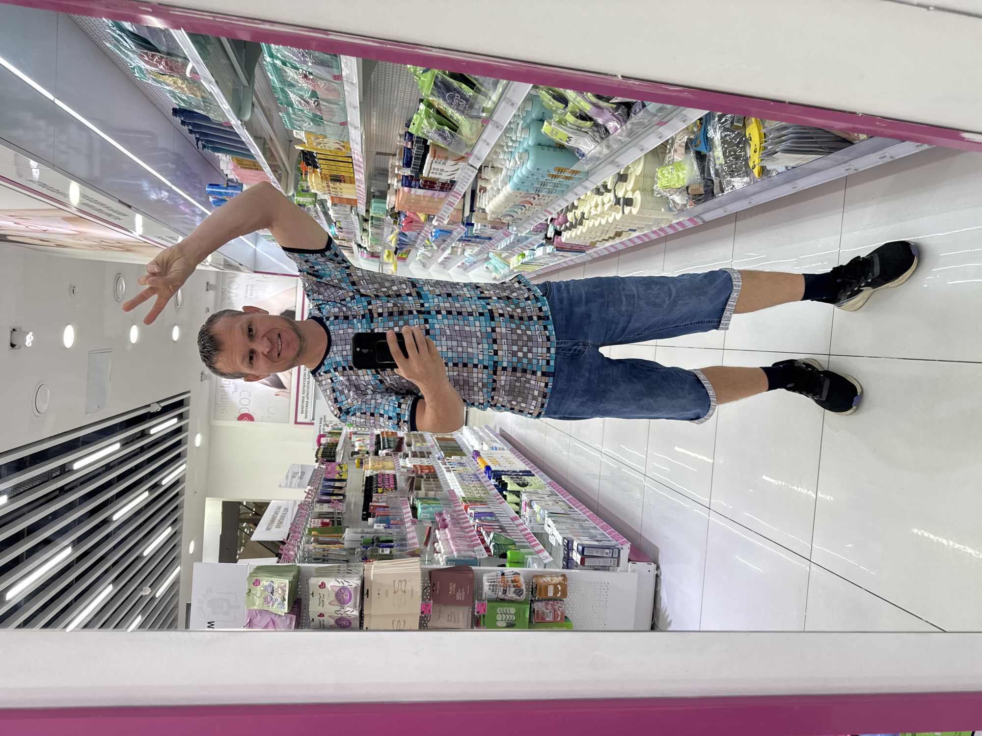 A man is reflected in a mirror in a store, likely a pharmacy or beauty supply store, in Oskemen, Kazakhstan. He is wearing a blue and white patterned t-shirt, denim shorts, black socks, and black athletic shoes. He is holding a black smartphone in his left hand and holding up two fingers in a peace sign with his right hand. The background shows aisles of shelves stocked with various products, including bottles of shampoo and lotion, and packaged beauty items. The floor is white tile, and the ceiling has exposed fluorescent lighting.