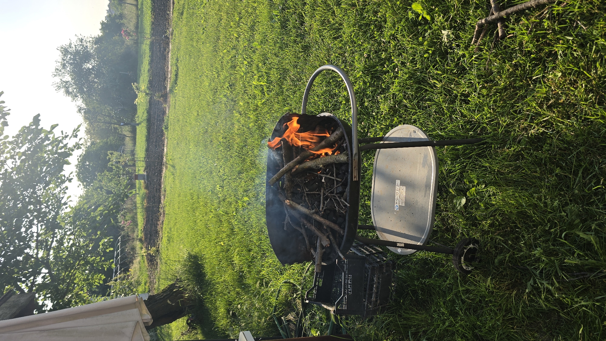 The scene depicts an outdoor garden or yard in the city of Rudnik, Bulgaria, under bright, low-angle sunlight, casting long shadows. In the foreground, a black kettle-style charcoal grill stands on a three-legged wheeled frame. Inside the grill, a fire burns with orange flames and light smoke, fueled by charcoal and numerous wooden branches and twigs. To the immediate left of the grill, two black plastic crates are stacked. A coiled green garden hose is partially visible behind these crates. The ground is covered predominantly by green grass, with some taller patches and scattered debris, including a few fallen branches to the lower right. In the middle ground, a cultivated plot of dark soil contains small plants or saplings. Within this cultivated area, a cylindrical object with red and white sections, possibly a well pump or planter, is visible towards the left. The background is characterized by dense green foliage from various trees and shrubs, with a bright light source (the sun) positioned high in the frame, creating lens flare effects. A light-colored fabric, likely from an umbrella or awning, is partially visible in the upper left corner, attached to a tree trunk.