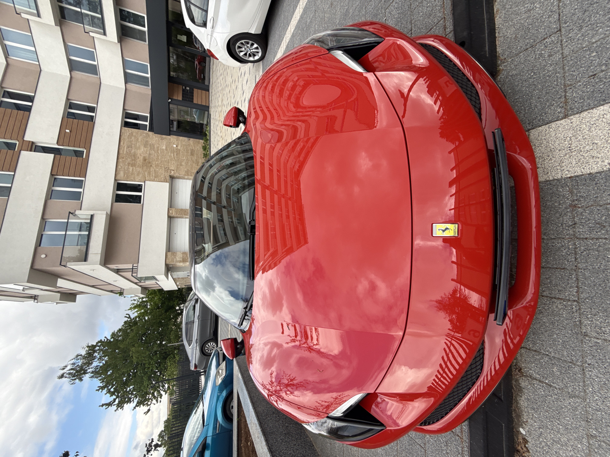 A bright red Ferrari faces the camera on a gray concrete pavement with a black stripe, reflecting the surroundings. In the background is a modern multi-story building with beige and white paneling and multiple windows. To the left, there's a view of greenery, including trees and a glimpse of a blue car and a silver car partially visible behind the Ferrari. The buildings and sky reflect clearly on the car's hood. This photo was taken in the city of Sofia, Bulgaria.