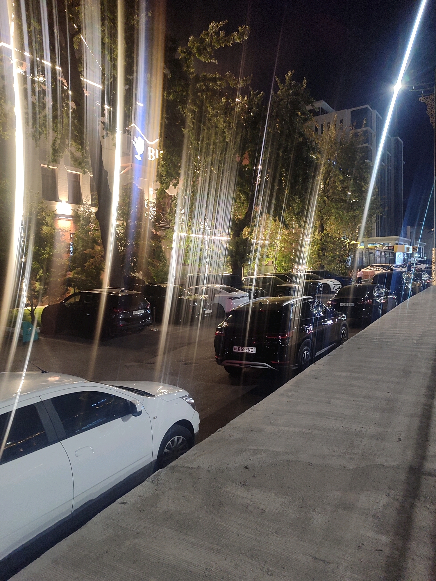 This image captures a night scene on a street in Tashkent, Uzbekistan. A row of parked cars lines the right side of the street, with a white car prominently featured in the foreground on the left. The street is illuminated by artificial lights, including what appear to be light installations that create vertical streaks of light, possibly from a water feature or light show. In the background, buildings and trees are visible. The overall impression is a lively urban setting at night.