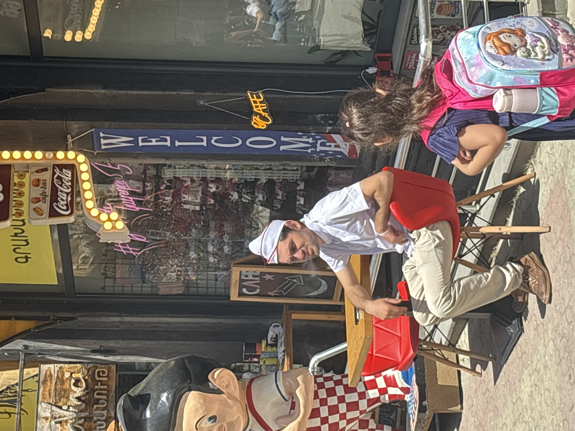 A man dressed as a diner worker, wearing a white shirt, hat, and red apron, is seated at a table outdoors. He appears to be interacting with something or someone to his right, his gaze directed downwards. A young girl with a pink and blue backpack stands to his right, her back mostly to the camera, looking in the same general direction as the man.
The scene is set in front of a building with large windows that reflect the sky and surrounding environment. A bright yellow neon sign shaped like a marquis with lights spelling out "cafe-bar" and "Coca-Cola" is visible on the left. A blue banner with "WELCOME" written in white letters hangs above, partially obscured by a small orange neon "CAFE" sign. Another wooden sign with "CAFE" written in white letters is propped against the building.
In the foreground, large cartoon-like figures, possibly mascots or decorations for the establishment, are visible. One is a character with black hair, wearing a white shirt with a red trim and a blue stripe. Another character is partially visible to the right of the first. A checkered red and white cloth is spread across part of the table.
The lighting suggests it is daytime, with bright sunlight casting shadows. The overall impression is that of an outdoor dining area or a street-side cafe, possibly in a tourist area, with a retro or diner theme.