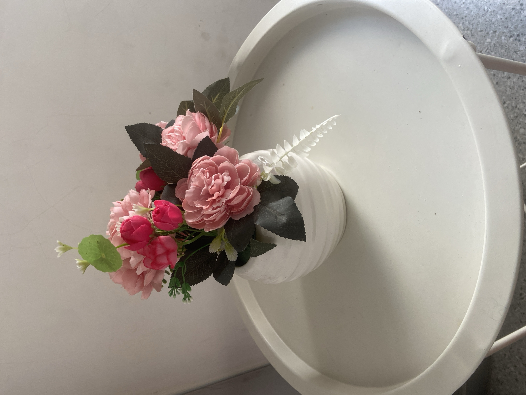 A close-up, eye-level shot shows a round, white ceramic vase filled with an arrangement of artificial pink and red flowers and dark green leaves. The vase is placed on a round, white table with a slightly raised edge. The background is a plain, light-colored wall. The scene is indoors and appears to be a still life composition. The image does not contain any information that can be used to determine its location in Tulungagung, Indonesia.