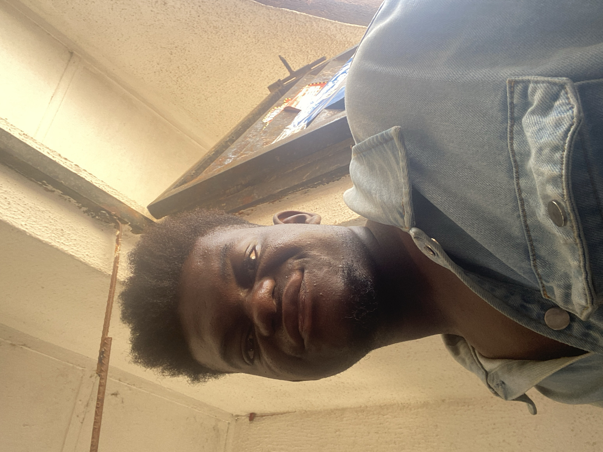 The image is an upward-angled shot of a dark-skinned male wearing a light blue denim jacket. He is looking down at the camera. The background includes a textured white or beige wall and a metal structure, possibly a beam or frame, with some debris or objects on top. The setting appears to be indoors. The location is Unknown, Unknown.