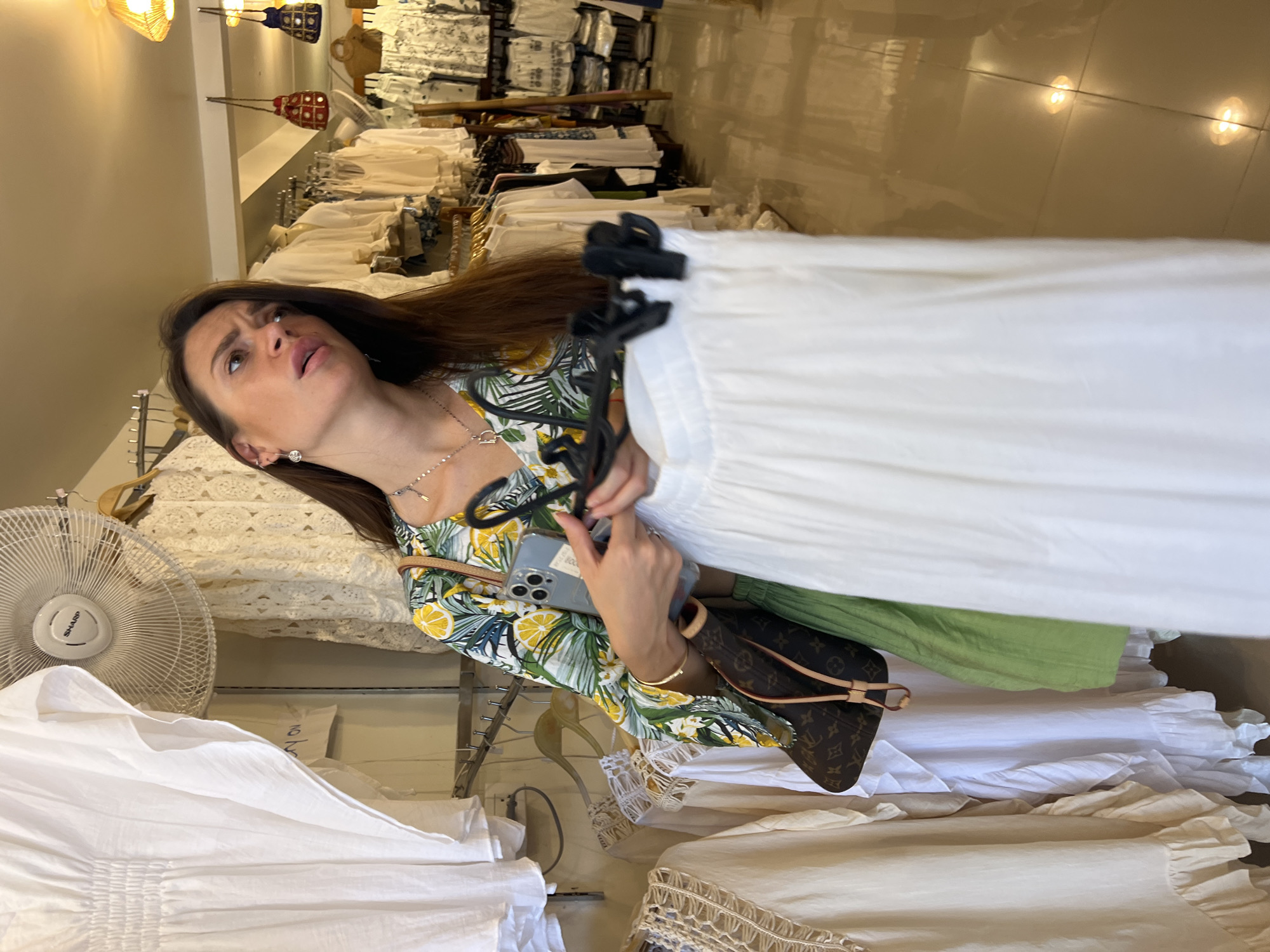 The photo shows a woman in a clothing store. She is holding a white dress on a hanger, looking up and to the left. She is wearing a printed floral dress, silver necklace, and gold bracelet. Her long brown hair is partly obscuring her view. She is holding a smartphone and a brown patterned handbag. The store is filled with racks of white clothing and a standing fan. The location is a clothing store in Baan Plai Laem, Thailand. The floor is tiled and reflective.
