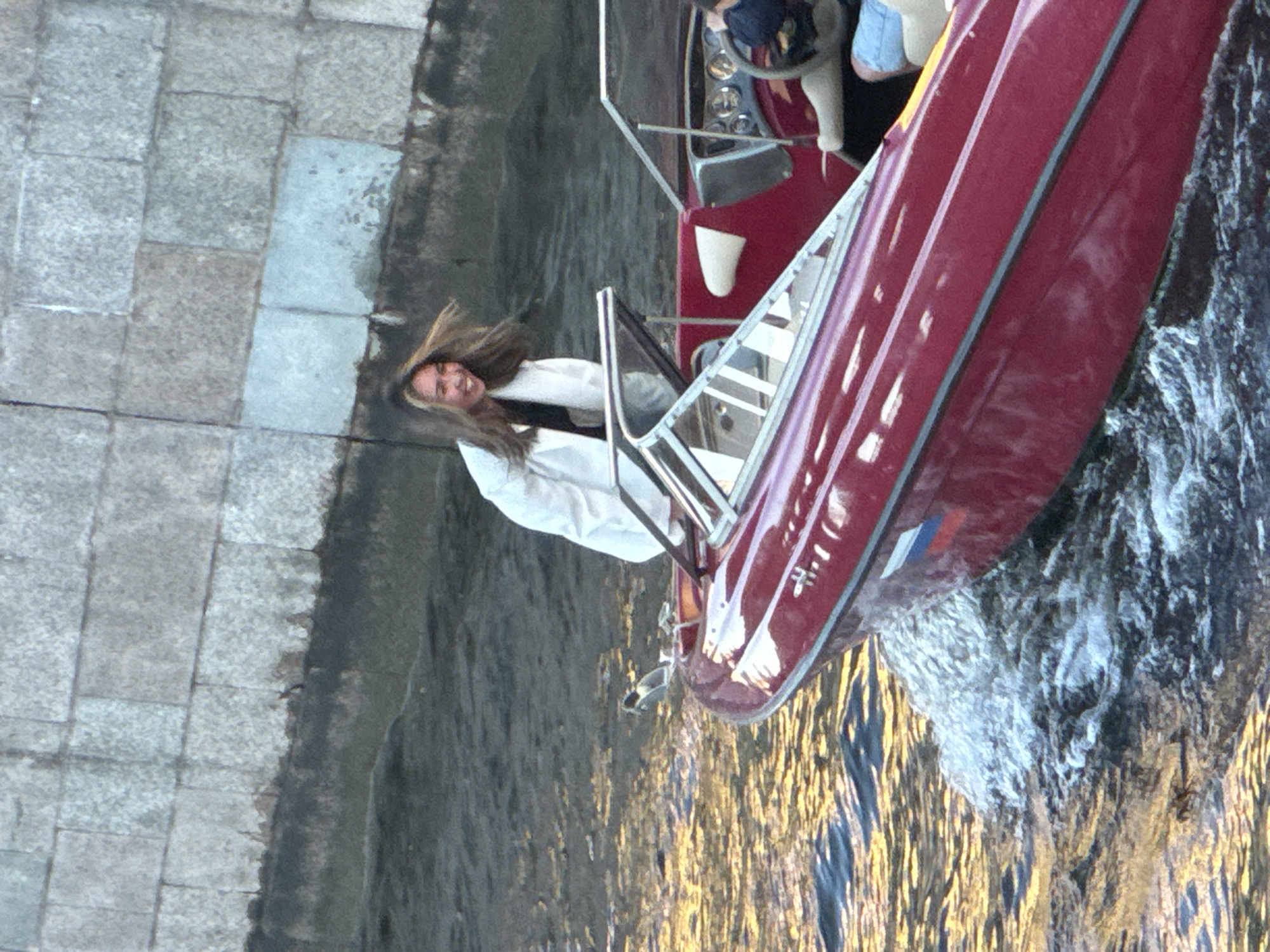 In this medium shot image, a young woman with long, wind-blown brown hair is standing on a red motorboat in Saint Petersburg, Russia. She is wearing a white jacket over a dark top and smiling joyfully at the camera. The boat is moving through dark water, leaving a wake. In the background, a stone wall of a building, likely along one of the city's canals or rivers, is visible. The sun is reflecting on the water, creating streaks of gold. Another person is partially visible in the boat, seated behind the steering wheel, wearing a dark top and blue shorts. The scene captures a moment of exhilaration on the water in Saint Petersburg.