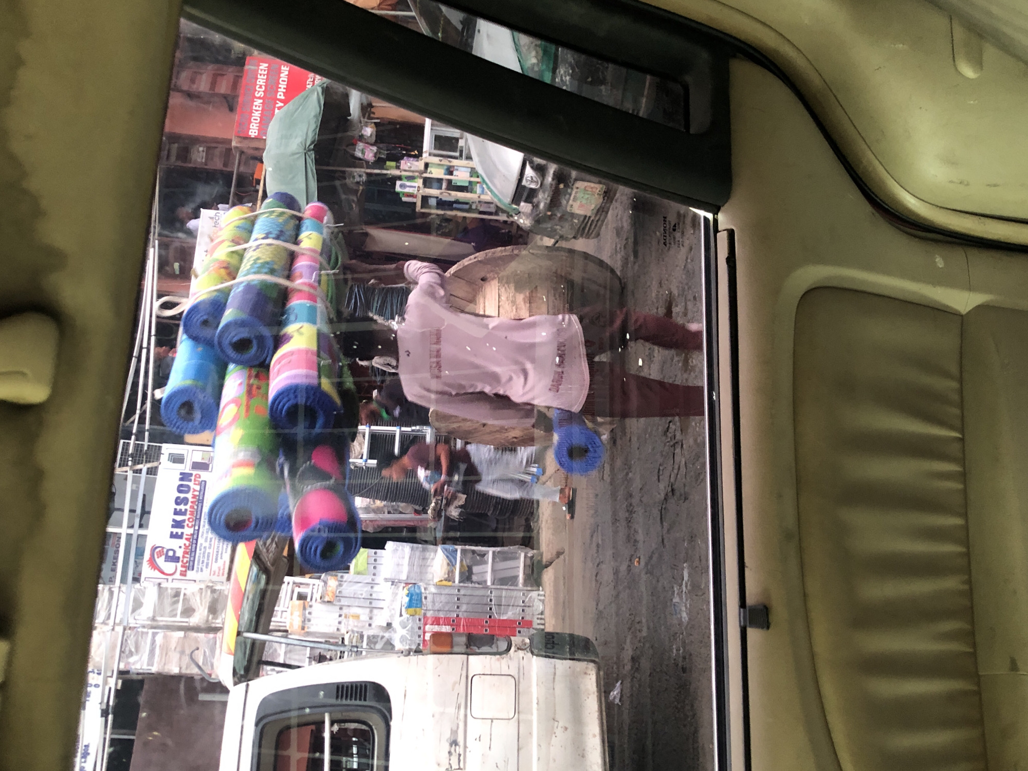 In Igbede, Nigeria, a person is seen from behind, wearing a pink long-sleeved shirt and maroon trousers, carrying a large bundle of colorful rolled-up mats balanced on their head and shoulder. The mats are tied together with rope. To the left, a white van is parked, its side visible. In the background, there are buildings with signs, including one that reads "P. EKESON ELECTRICAL COMPANY LTD". Various goods are displayed on shelves and stalls, suggesting a marketplace or commercial area. A silver car is parked to the right of the scene. The ground appears to be dirt and somewhat muddy.
