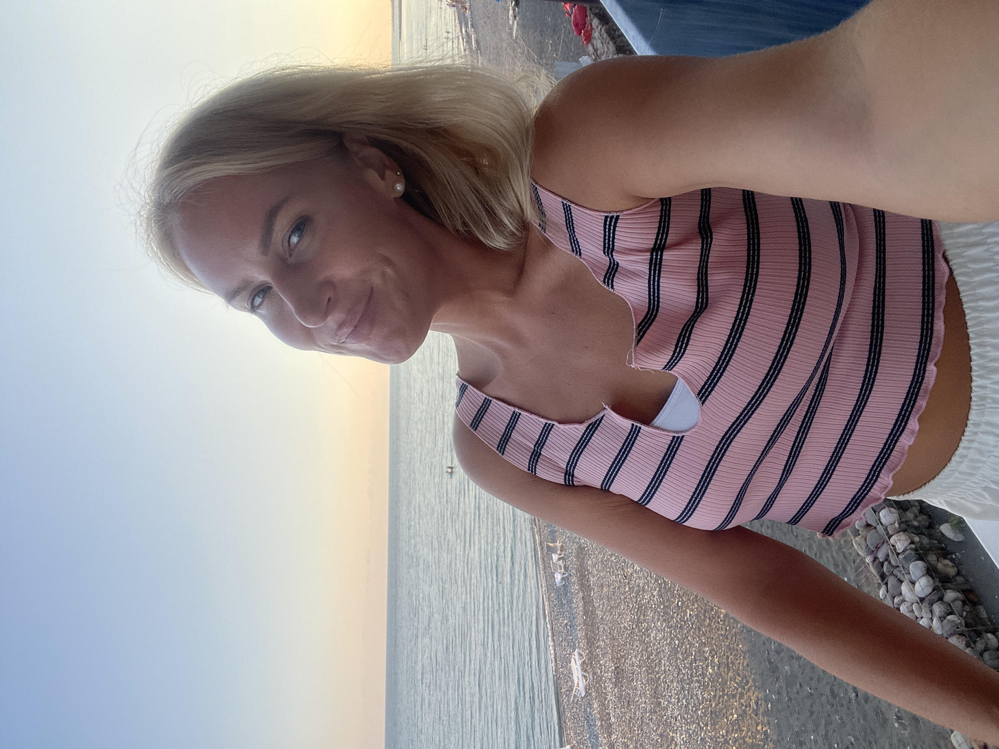 A woman with blonde hair and blue eyes, wearing a pink and black striped top and white shorts, smiles at the camera while taking a selfie. She stands on a balcony overlooking a pebble beach and the sea. The sky is a pale blue and yellow, suggesting it is either sunrise or sunset. There are a few beach chairs on the shore, and some people are visible in the water. The location is identified as Giachrypsh, Abkhazia.