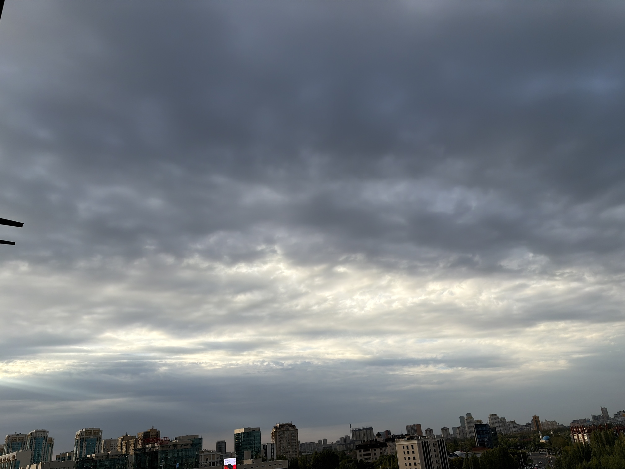 The image depicts a wide-angle, slightly tilted view of a cityscape under a cloudy sky. The scene is dominated by various buildings of different heights and architectural styles, ranging from modern high-rises to older, more ornate structures. Trees and greenery are visible interspersed between the buildings, suggesting an urban environment with parks or tree-lined streets. The sky is heavily overcast with dark, textured clouds, but there are areas where sunlight is breaking through, creating brighter patches and beams of light. The overall lighting suggests it could be late afternoon or early evening. The location is identified as Astana, Kazakhstan. There are no people or explicit actions visible in the image, making it a static, environmental shot.