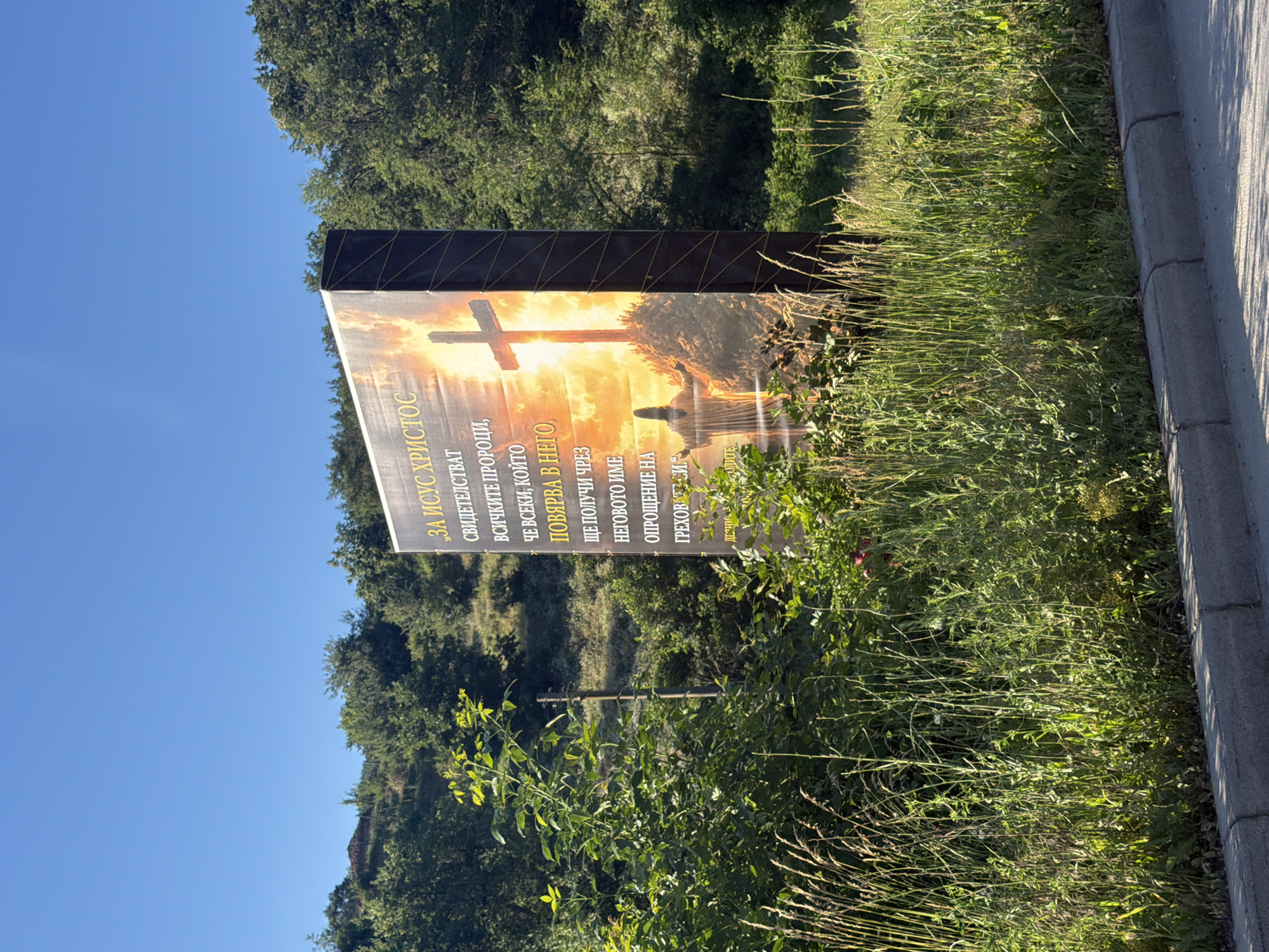 A large, rectangular billboard featuring a religious image is prominently displayed outdoors. The billboard is positioned at an angle, with its right side closer to the viewer. The image on the billboard depicts a sunset or sunrise over a hill, with a large cross silhouetted against the bright sky. A figure of Jesus Christ, arms outstretched, is visible at the base of the hill. Text is present on the billboard in what appears to be Cyrillic script.
The billboard is surrounded by lush green vegetation, including tall grasses and bushes. The foreground shows a grey concrete curb and pavement, suggesting a roadside location. In the background, there are trees and a hillside, characteristic of a rural or semi-rural environment. The sky is clear and blue, indicating fair weather.
The scene is located in the city of Veselinovo, Bulgaria, as indicated by the context provided. The overall impression is one of a roadside religious advertisement set against a natural landscape.