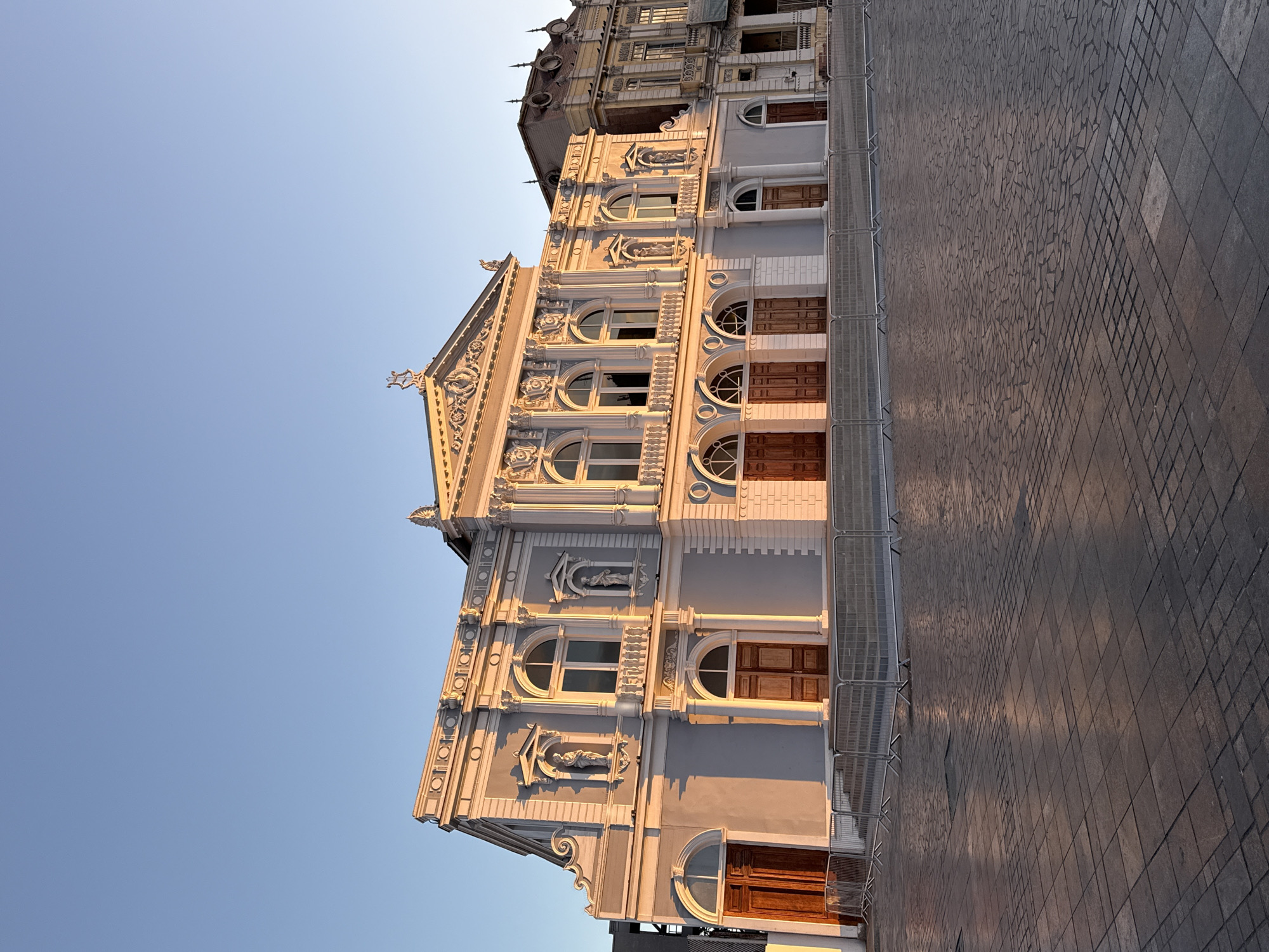 The image depicts the Teatro Municipal de Iquique in Iquique, Chile, during what appears to be sunset or sunrise, given the warm, low-angle lighting casting long shadows and highlighting the architectural details. The building is a grand, neoclassical structure painted in a light blue-grey with white decorative elements. It features multiple arched doorways and windows, balconies, and sculpted figures adorning the facade. The building is flanked by another, older-looking structure on the right, which is yellow and shows signs of wear. In front of the building, a plaza paved with intricate stone patterns is visible. Metal barriers are set up, suggesting a cordoned-off area. The sky is a clear, pale blue, with no clouds present. There are no people or vehicles visible in the scene.