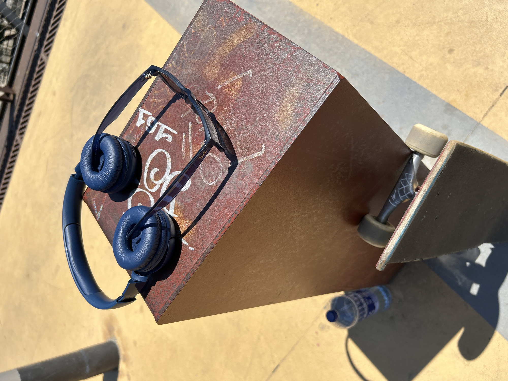 The scene is an outdoor setting, likely a skate park, located in the city of L'Hospitalet de Llobregat, Spain. The ground is primarily yellow with some grey concrete sections. A rusted metal object, perhaps a skate park feature, is centrally located in the frame. On top of this metal structure, there are blue wireless headphones and a pair of sunglasses. A skateboard with one visible white wheel is leaning against the metal object. A partially visible water bottle sits near the base of the metal structure. The lighting indicates a sunny day with distinct shadows cast by the objects. There are no people visible in the image. Graffiti-like markings are present on the metal object.