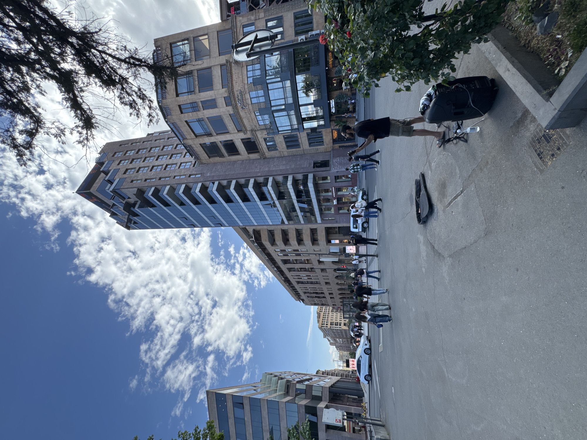 The image shows a street scene in Yerevan, Armenia. Several modern buildings of various heights line the street. A street musician with a guitar stands next to an amplifier. A few pedestrians are walking on the sidewalk. A white car is parked on the right side of the road. The sky is blue with scattered clouds.