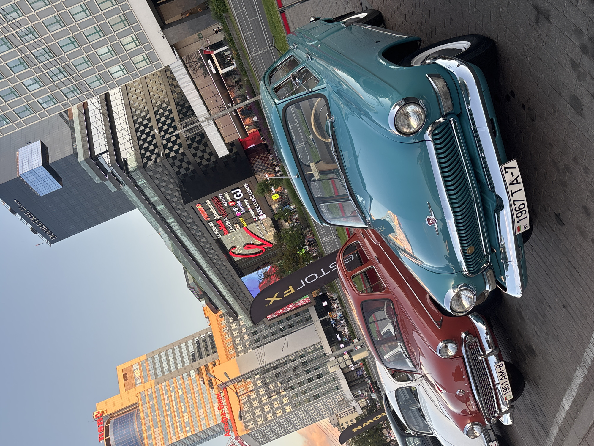 In the city of Unknown, Unknown, on a paved surface with a brick pattern, several vintage cars are parked in a row. The closest car, a turquoise sedan, is prominently featured in the foreground. It has a chrome grille, round headlights, and a license plate with the text "BY 1967 TA-7". Behind it, a red sedan with a similar design and a license plate reading "BY 1961 AM-8" is visible. Further back, a white car and a black car are partially visible. In the background, modern buildings rise, some with glass facades and distinct architectural designs. One building displays a large digital screen with colorful advertisements, and other signs indicate retail establishments like "LC Waikiki," "Stradivarius," and "KFC." The sky is clear and bright, suggesting daylight. The overall scene captures a juxtaposition of vintage automotive design against a backdrop of contemporary urban architecture.
