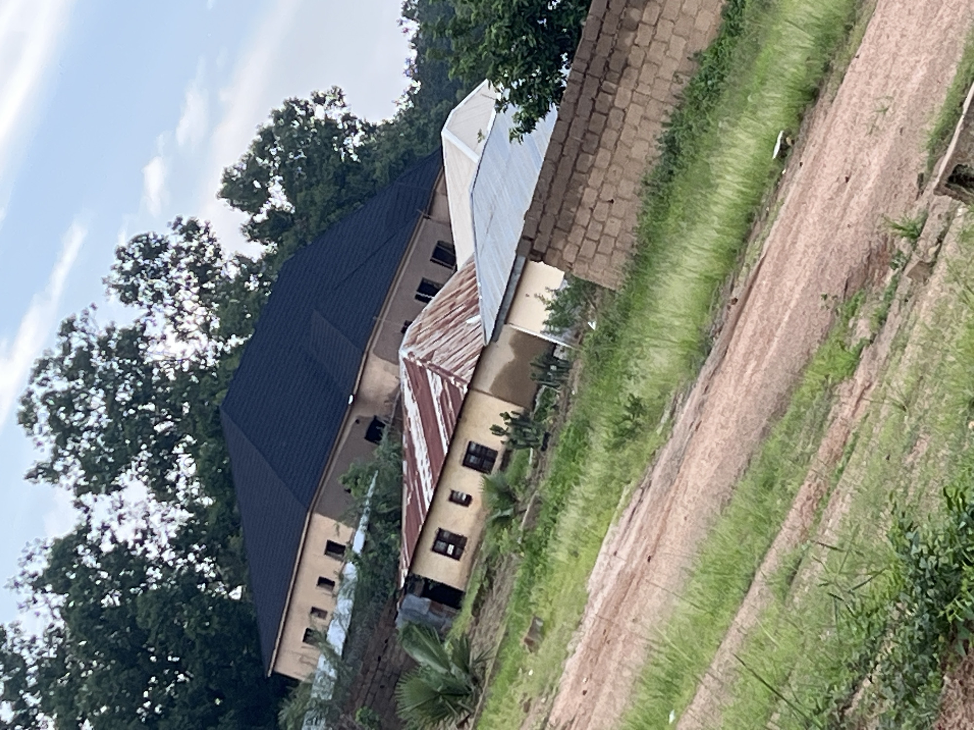 The image depicts a residential area in Gura Top, Nigeria. In the foreground, a dirt path is visible, bordered by green grass. Further back, several structures are present. The most prominent building is a two-story house with a dark, sloped roof. To the left and slightly lower, another building with a weathered, corrugated metal roof is situated, featuring small windows. To the right of this, a lighter-colored structure with a pale roof is visible, and behind it, a wall constructed of tan bricks. The scene is surrounded by lush green trees, with a partly cloudy sky overhead. There are no people or vehicles visible in the image. The overall impression is of a developing or established residential neighborhood.
