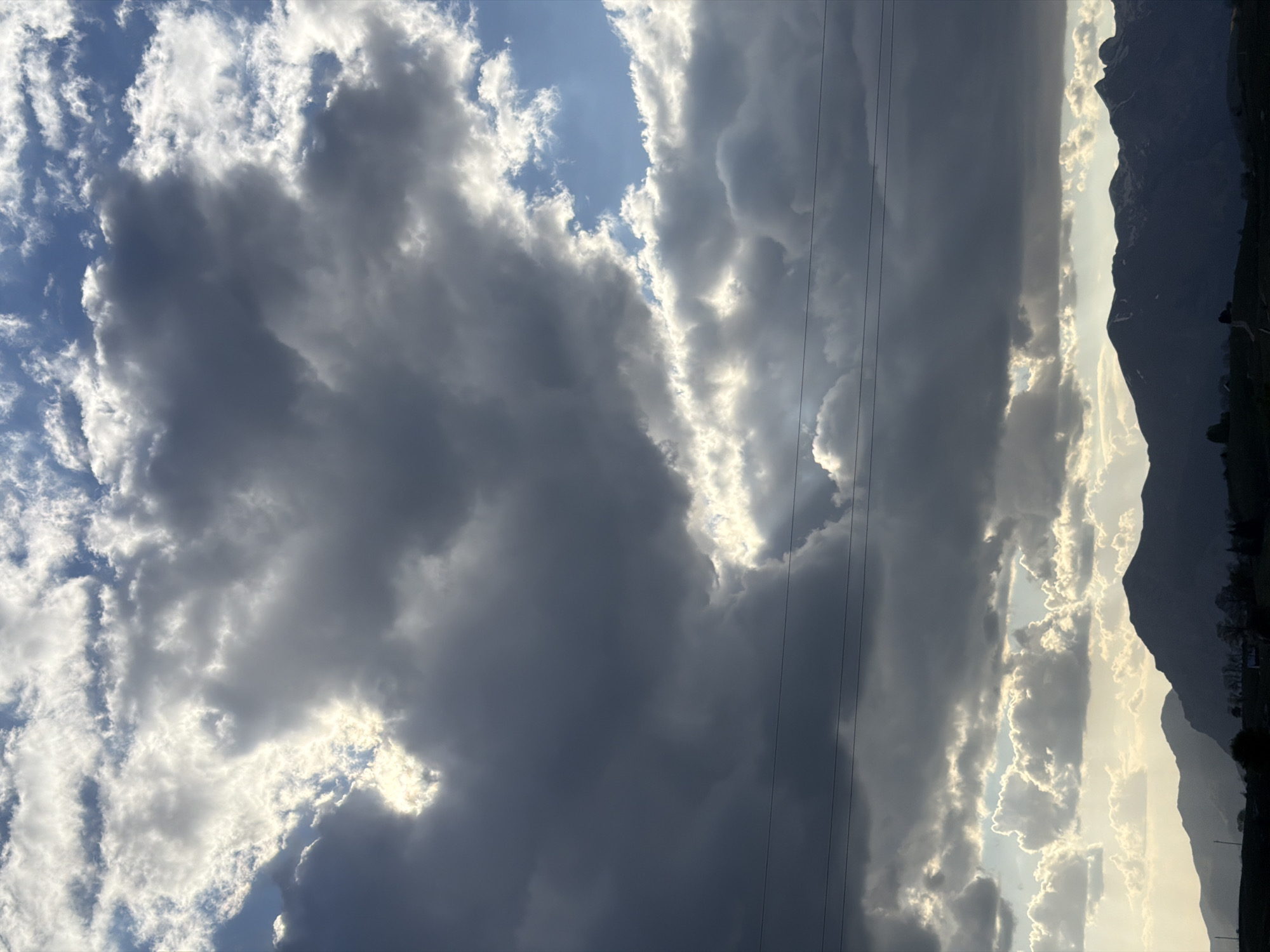 The image shows a sky filled with dark, heavy clouds interspersed with patches of sunlight. Mountains are visible on the right side of the frame, silhouetted against a brighter horizon. Thin power lines cut across the sky. The scene is of the city of Pišče, Montenegro.