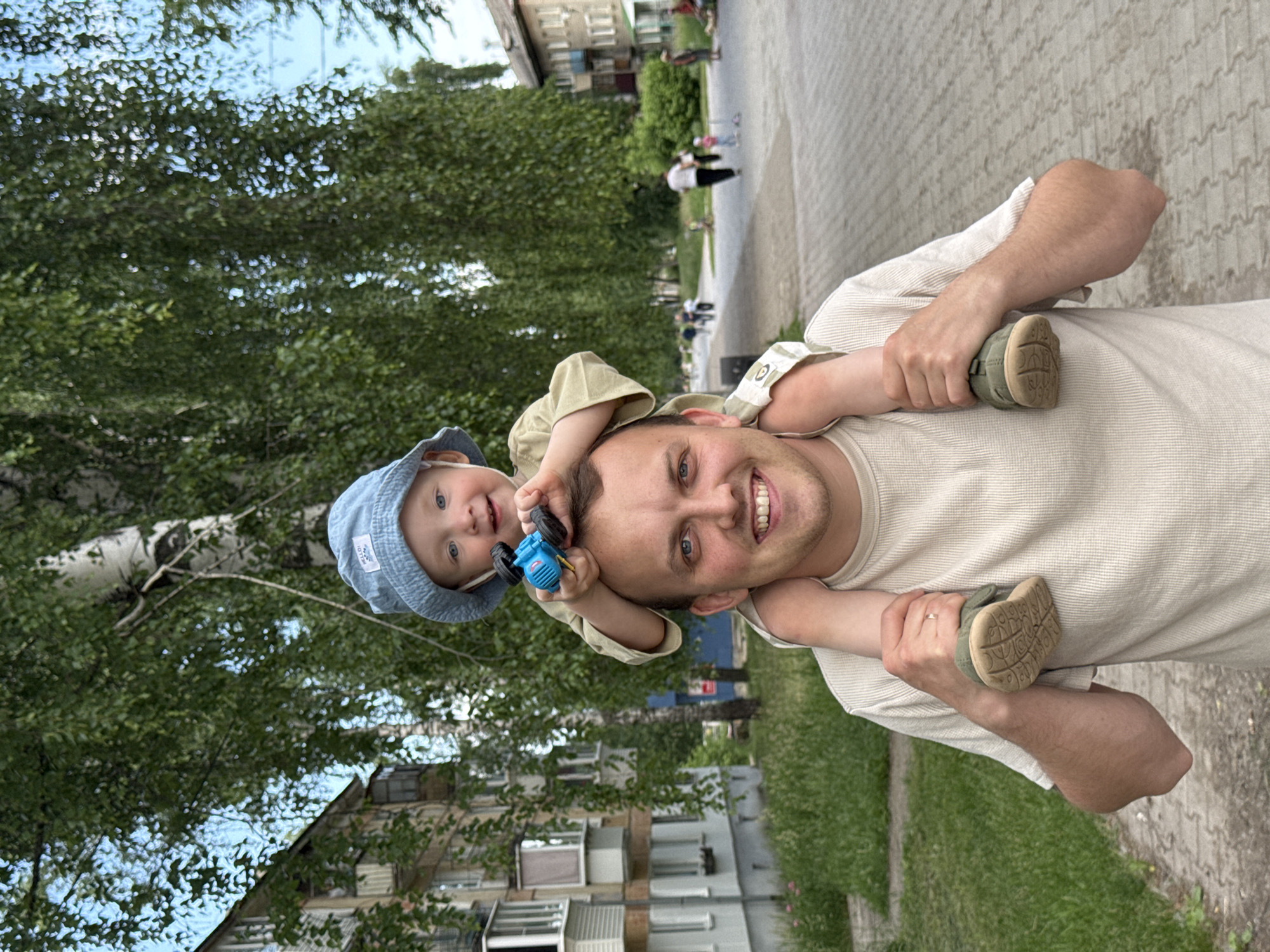 A father carries his young son on his shoulders outdoors, in a city park. The father, a Caucasian male with short brown hair and blue eyes, wears a beige waffle-knit t-shirt. His son, also Caucasian with blonde hair and blue eyes, is seated on his father's shoulders, wearing a light blue bucket hat and a beige onesie. The child holds a small blue toy car in his right hand, which he is offering to his father.
The background shows a paved walkway lined with green grass and trees. Beyond the walkway, residential buildings are visible. In the distance, several people are walking, suggesting this is a public park in an urban setting. The sky is partly cloudy. The overall impression is a casual, happy moment between father and son. The presence of apartment buildings and the type of trees suggest this location could be in Chaikovsky, Russia, a city known for its parks and residential areas.