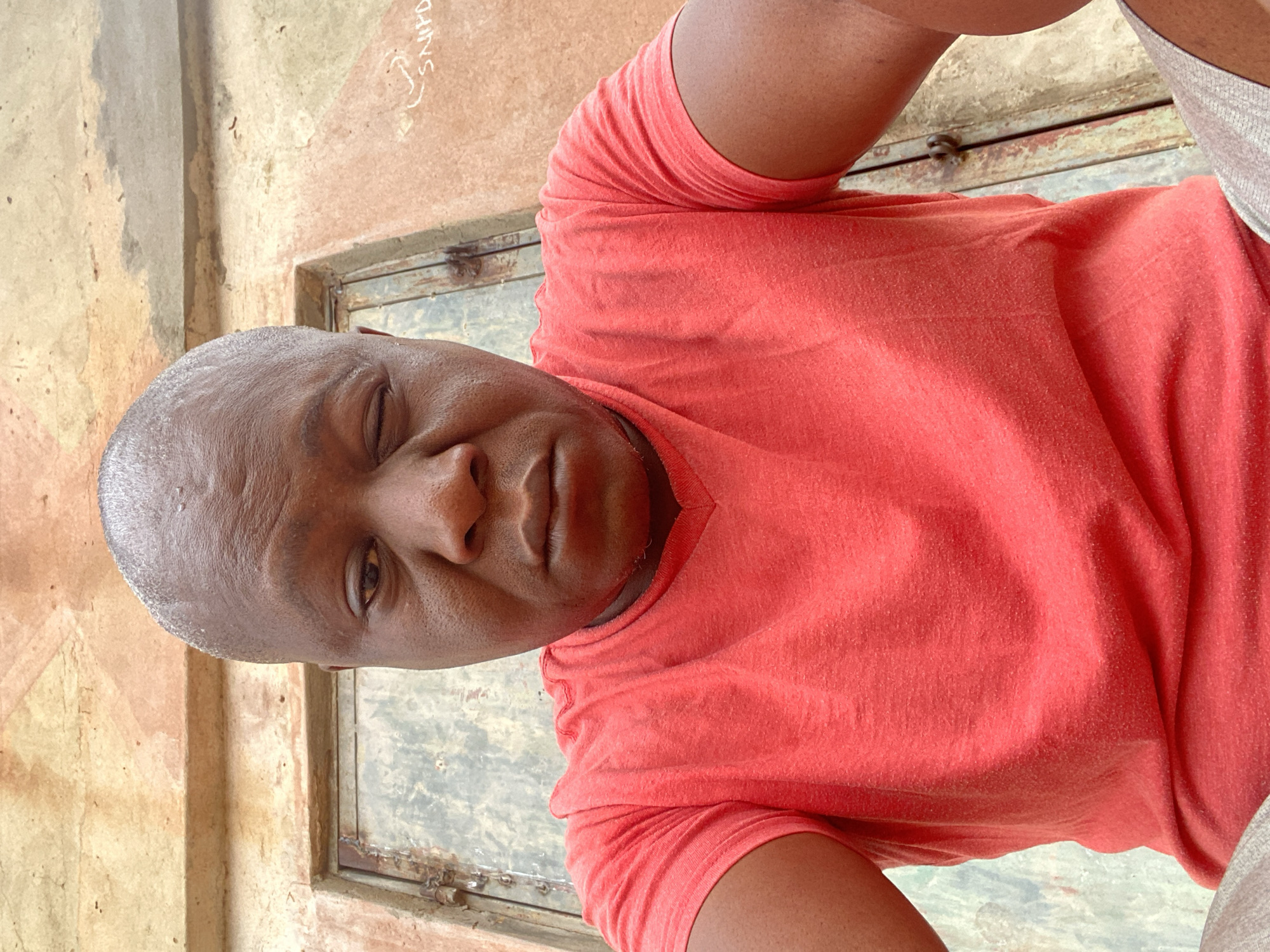 A close-up, waist-up shot shows a dark-skinned man with a shaved head. He is wearing a reddish-orange t-shirt. His face is angled slightly to the side, with one eye partially closed and a neutral expression. The background consists of a weathered wall with a small, barred window. Based on the metadata, the location is Sabon Tasha, Nigeria. No actions or interactions are visible. The overall environment appears to be outdoors in a run-down setting.