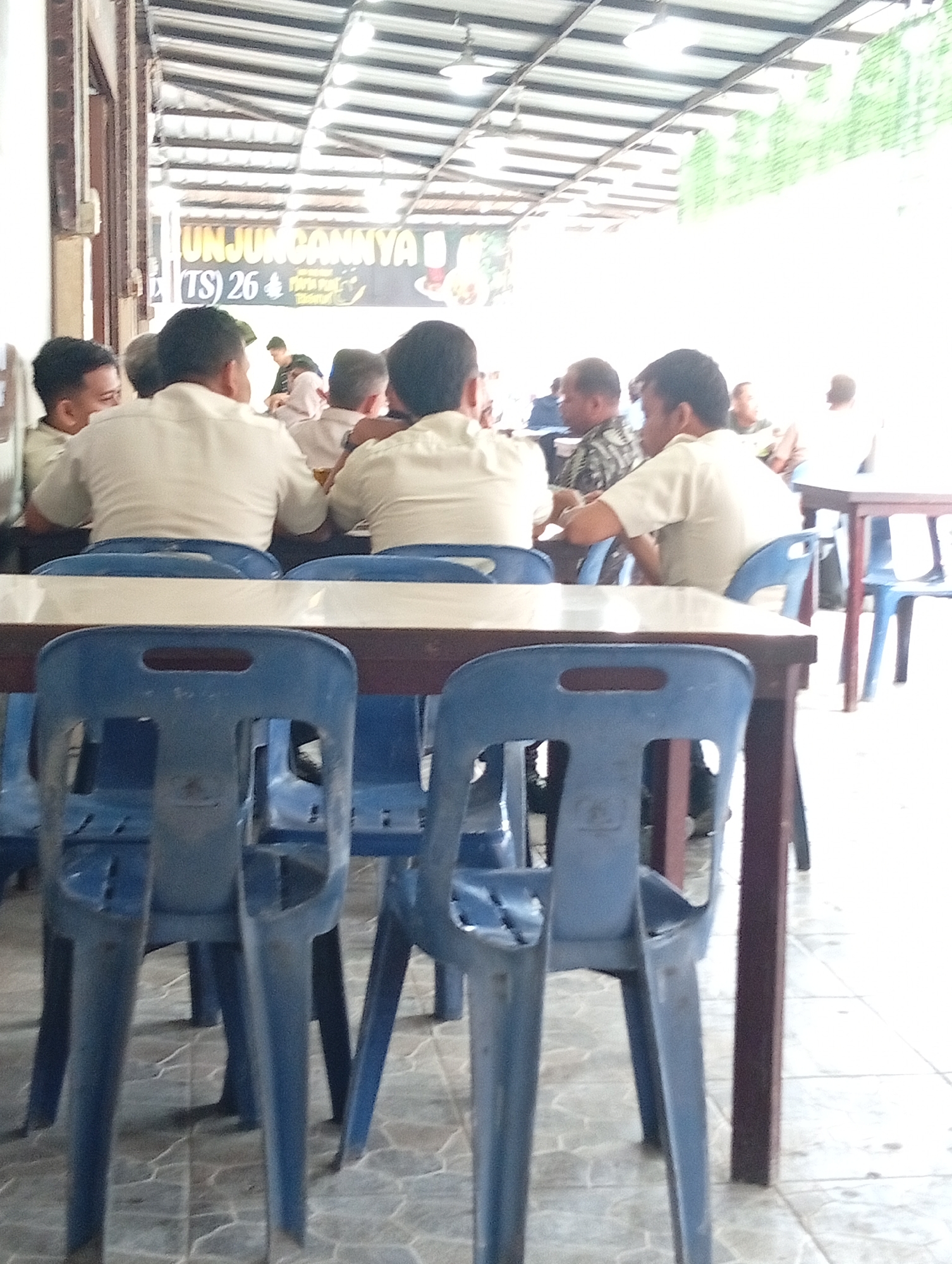 This media file depicts an informal gathering of men in an indoor-outdoor setting, likely a casual restaurant or meeting area in Lhokseumawe, Indonesia. The bright natural lighting suggests it is daytime.
The scene features numerous adult men, mostly viewed from behind or in profile. Many are wearing light-colored, possibly cream or off-white, short-sleeved shirts, which could indicate a uniform or common dress code. They are seated around several wooden-topped tables, utilizing simple blue plastic chairs. In the foreground, a table and several blue plastic chairs are prominent. Further back, more men are visible, appearing engaged in conversation, a meeting, or dining. One man in the mid-ground wears a patterned shirt, distinguishing him from those in light-colored attire.
A large banner or sign is visible in the background, partially obscured by the people and the building structure. Clearly legible text on the banner includes "UNJUNGANNYA" and "(TS) 26". The venue features an open design with a corrugated metal roof, typical of establishments in warm climates. The floor is covered with light-colored patterned tiles. The overall atmosphere appears calm and focused, with individuals engaged with one another.