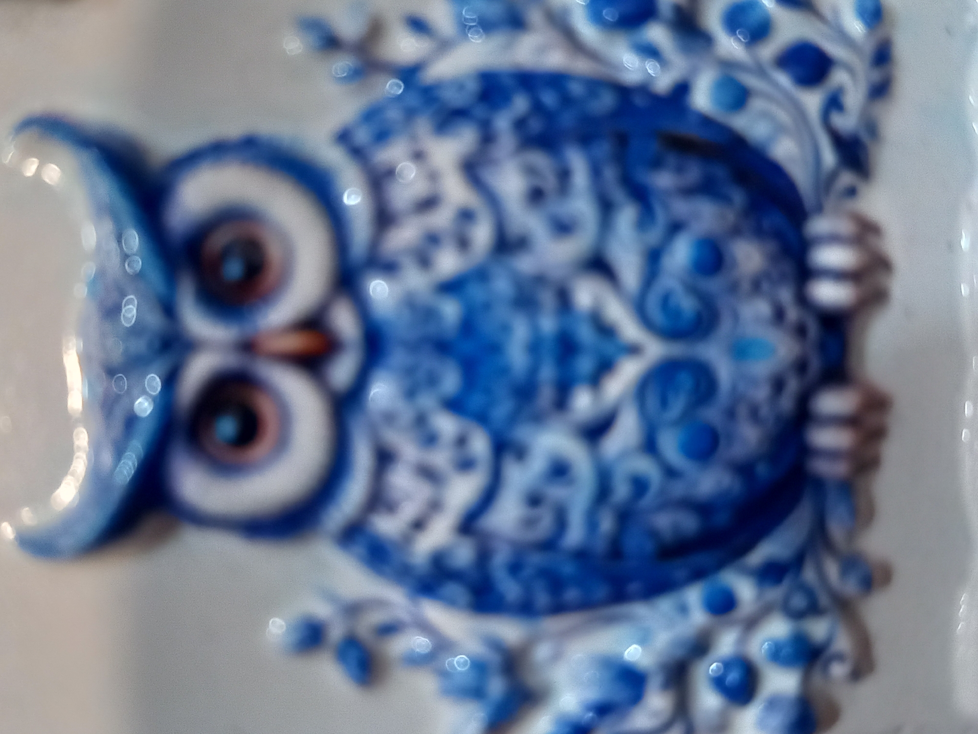 The media file depicts a close-up, blurred view of a decorative ceramic owl figurine. The owl is predominantly blue and white, featuring an ornate, traditional pattern reminiscent of Delftware or Gzhel ceramics. Its large, circular eyes are a focal point, with dark pupils surrounded by concentric rings of white and brown. The owl's body is adorned with swirling, decorative motifs and circular blue dots. Its wings are tucked in, and it appears to be clinging to something, with its hands or claws visible on the right side of the frame, also rendered in blue and white. The background is a soft, light gray, suggesting a neutral surface or wall. The overall impression is one of a static, decorative object, likely displayed indoors. The blurriness of the image, along with the bright, circular bokeh effects, suggests the photograph was taken with a shallow depth of field or in less than ideal lighting conditions, possibly with a flash. There is no visible text in the image.