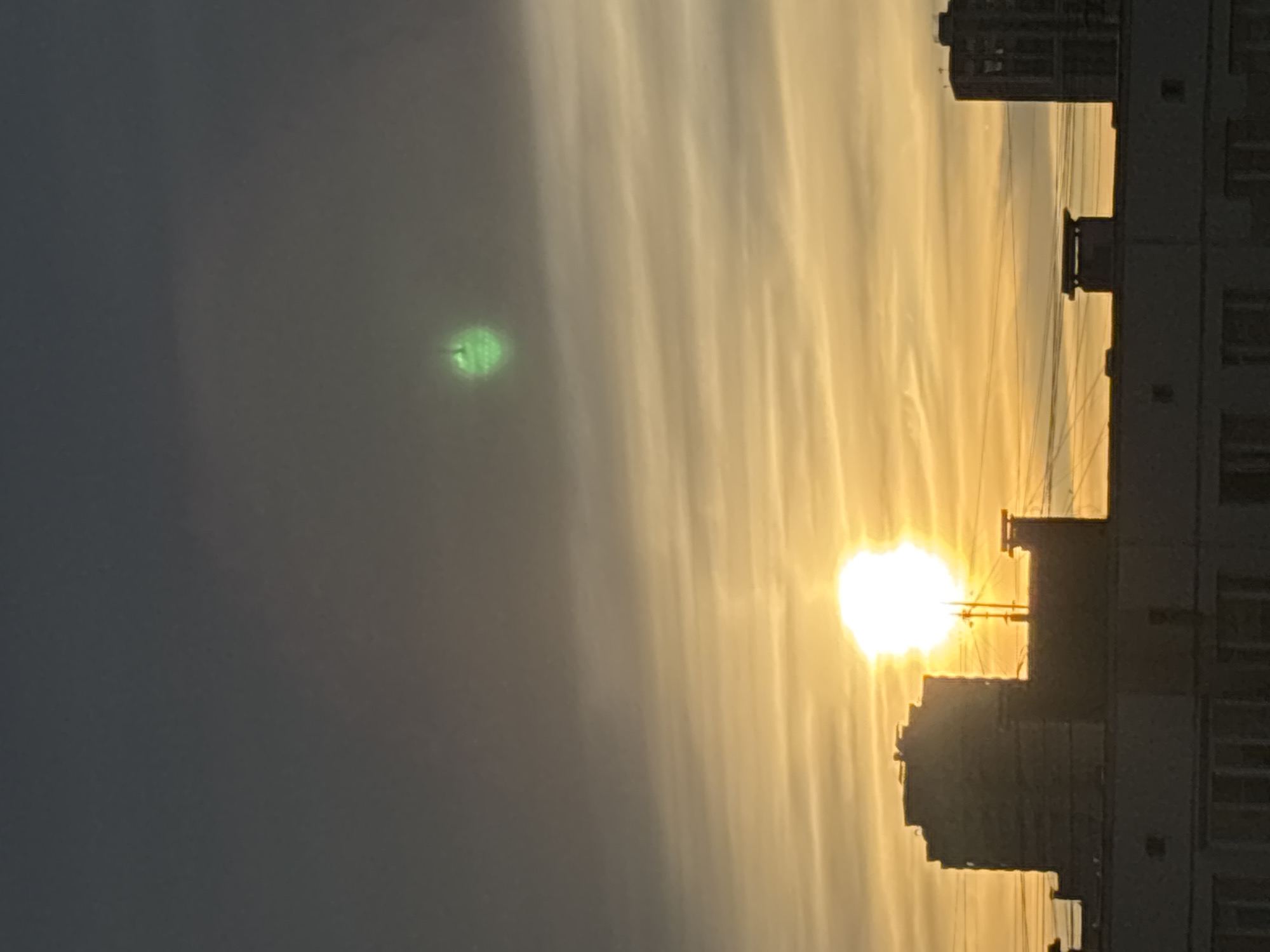 The image shows a sunset over an urban landscape in Unknown, Russia. The sun is partially obscured by buildings, casting a golden light across the sky with linear cloud formations. A bright, unidentified green orb is visible in the upper left quadrant of the image, within the darker section of the sky. Power lines are seen silhouetted against the sun. The focus is primarily on the interplay of light and shadow caused by the sunset and the buildings in the foreground.