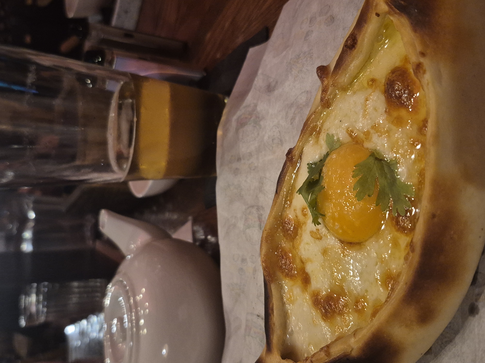 The media file displays a close-up view of a khachapuri, a traditional Georgian cheese-filled bread, placed on a white paper liner on a wooden surface. The khachapuri is boat-shaped, with a golden-brown crust. It is generously filled with melted white cheese, and in the center, a bright orange egg yolk rests, topped with a sprig of fresh green herb, likely cilantro. The cheese appears slightly browned and bubbly in places, indicating it has been freshly baked.
In the background, to the left of the khachapuri, is a glass bottle with a yellow liquid. Further behind that, a white ceramic teapot with a handle is visible, partially obscured by the bottle. The lighting suggests an indoor setting, possibly a restaurant, with warm tones dominating the scene. The orientation of the image is somewhat tilted. There is no visible text in the media file. The overall impression is of a hearty and appealing dish ready to be enjoyed.