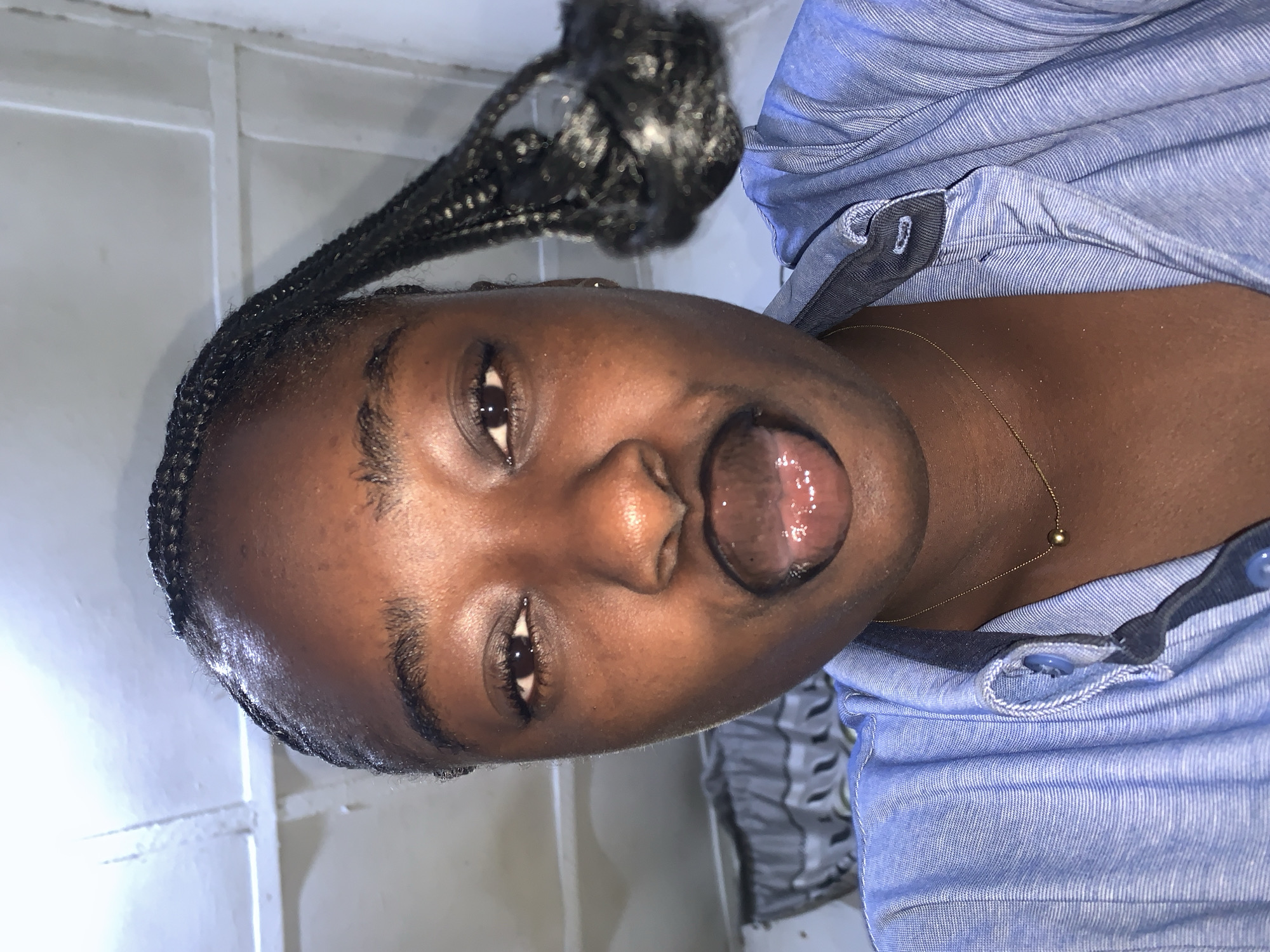 A young Black woman with braided hair is looking into a camera and sticking her tongue out. She is wearing a light blue button-down shirt and a simple gold necklace with a single bead. Her lips are glossy and appear to be painted black. The background is indistinct, but appears to be an interior space with white walls and some hanging fabric. The lighting suggests artificial light. The location is not visible or identifiable from the image.