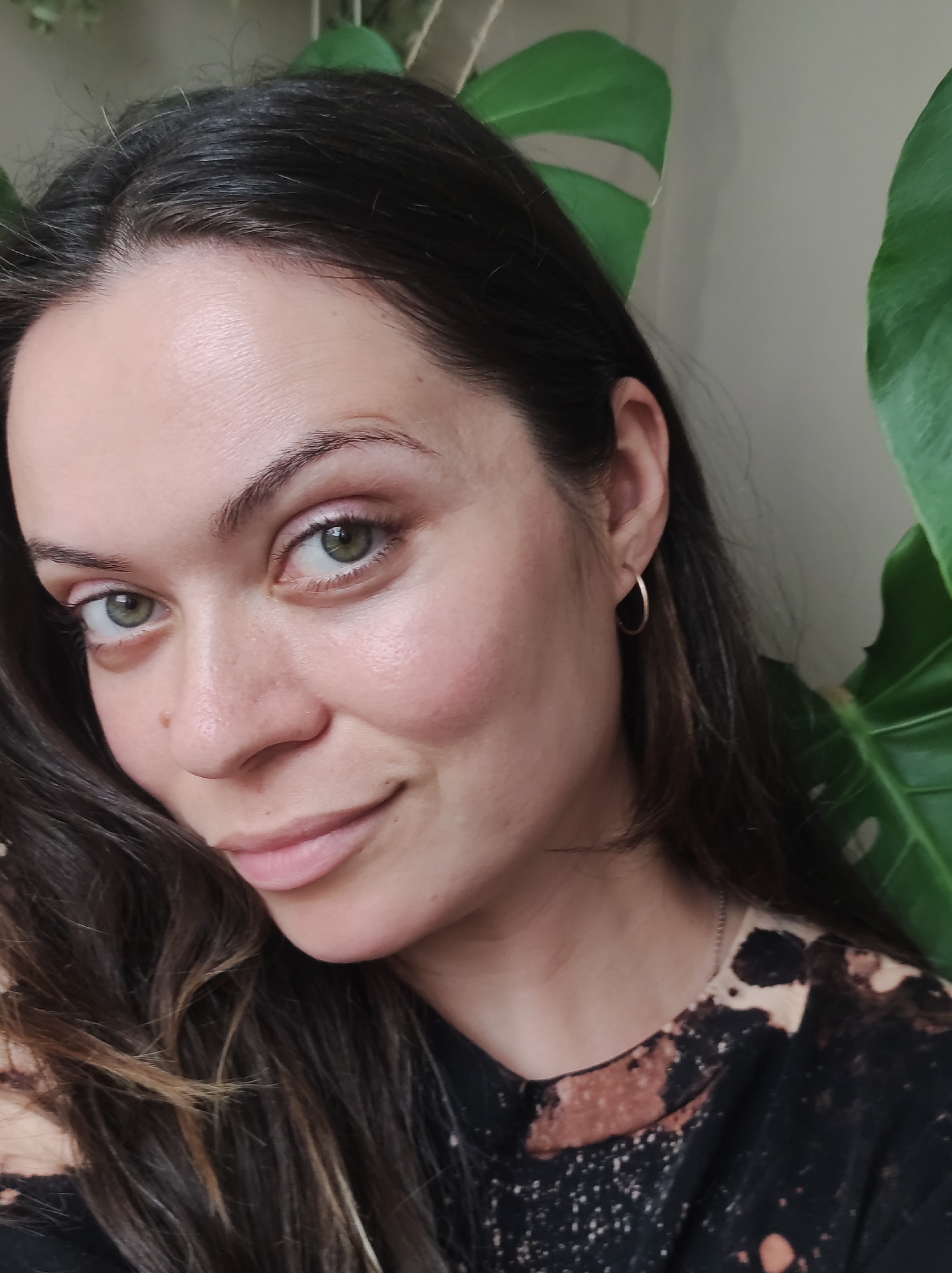 A close-up shot of a woman with fair skin, green eyes, and dark hair. She is wearing a small gold hoop earring in her left ear and a black t-shirt with a tie-dye pattern. Behind her, there is a large green plant with broad leaves. The background is a light neutral color. There is no indication of Chaikovsky, Russia in the image.