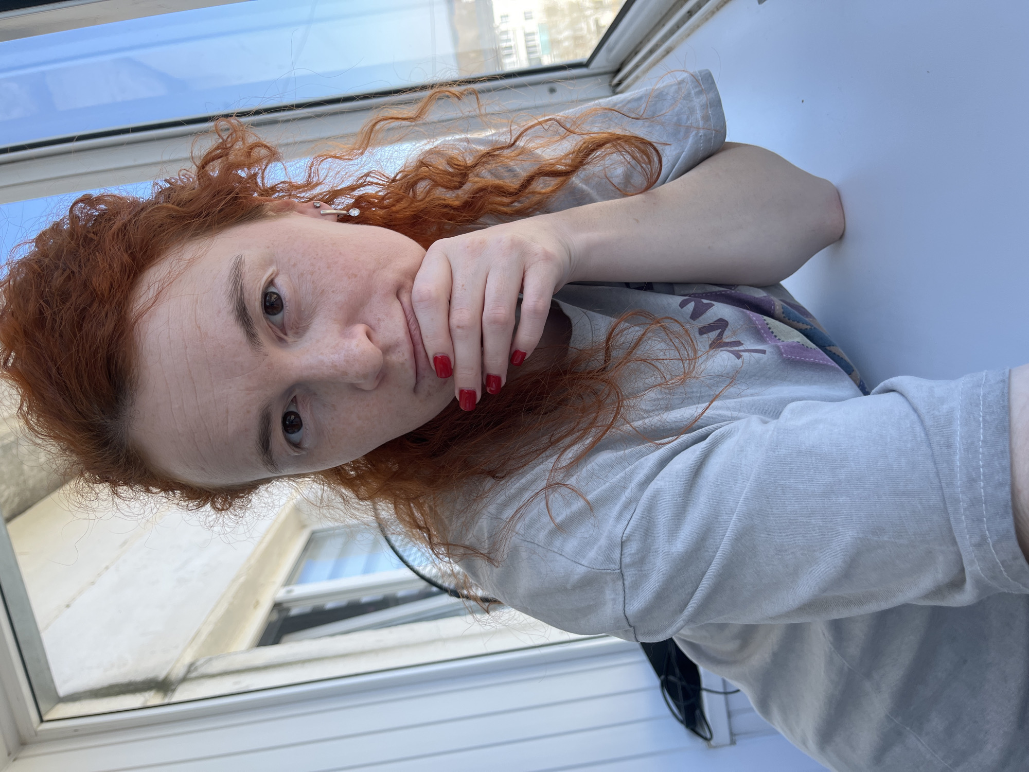 A woman with red hair and freckles is posing indoors in front of a window. She is wearing a light gray t-shirt and has red nail polish on her fingernails. Her hand is resting on her chin. Outside the window, a building is visible, suggesting an urban environment. Based on the prompt, the location is Chaikovsky, Russia.