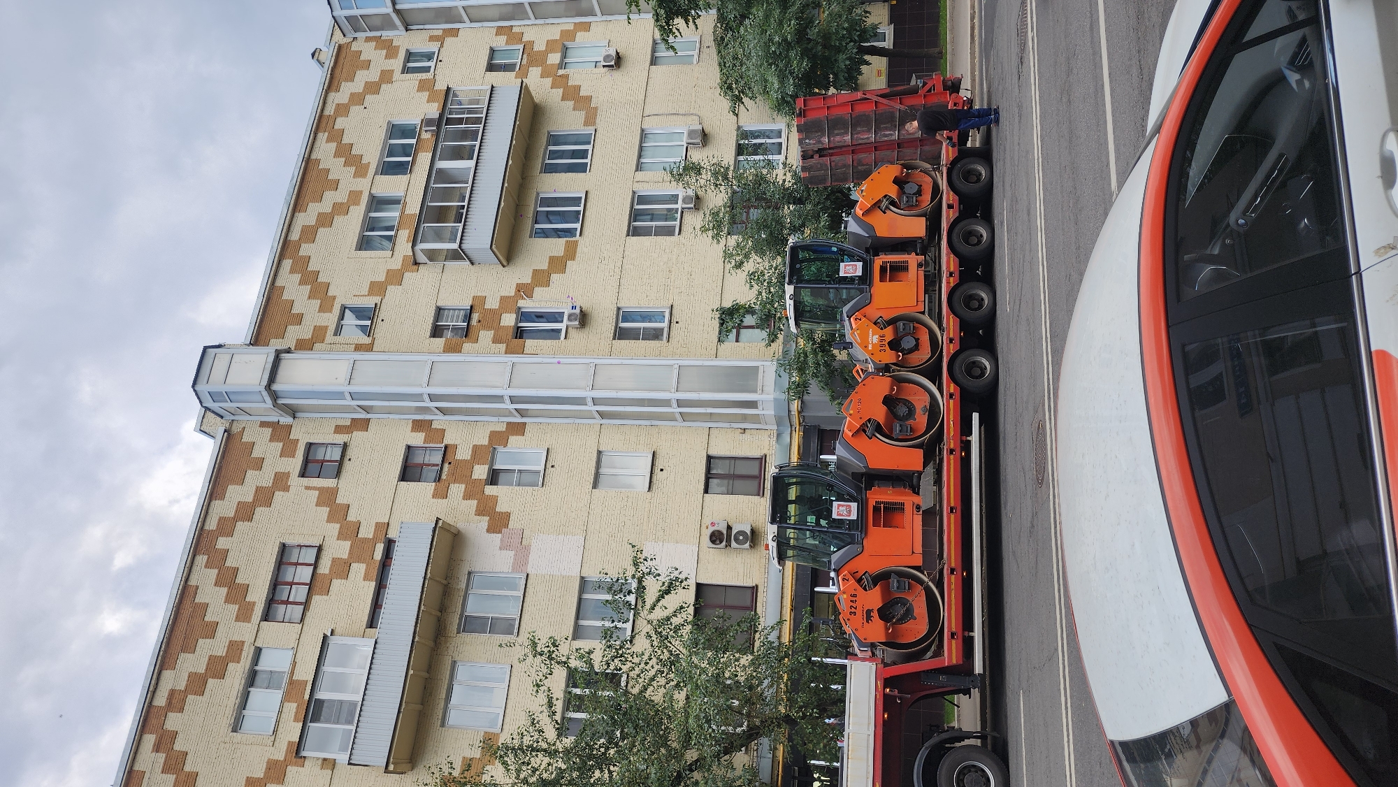 In Moscow, Russia, a flatbed truck is transporting two orange steamrollers on a city street. The truck is parked near a large, multi-story apartment building with a cream-colored facade decorated with a zigzag pattern of brown bricks. A glass-enclosed elevator shaft runs vertically up the side of the building. In the foreground, the roof of a white and orange car is visible. A man in a dark shirt and blue jeans stands at the rear of the truck, facing away from the camera. The sky is overcast with clouds.