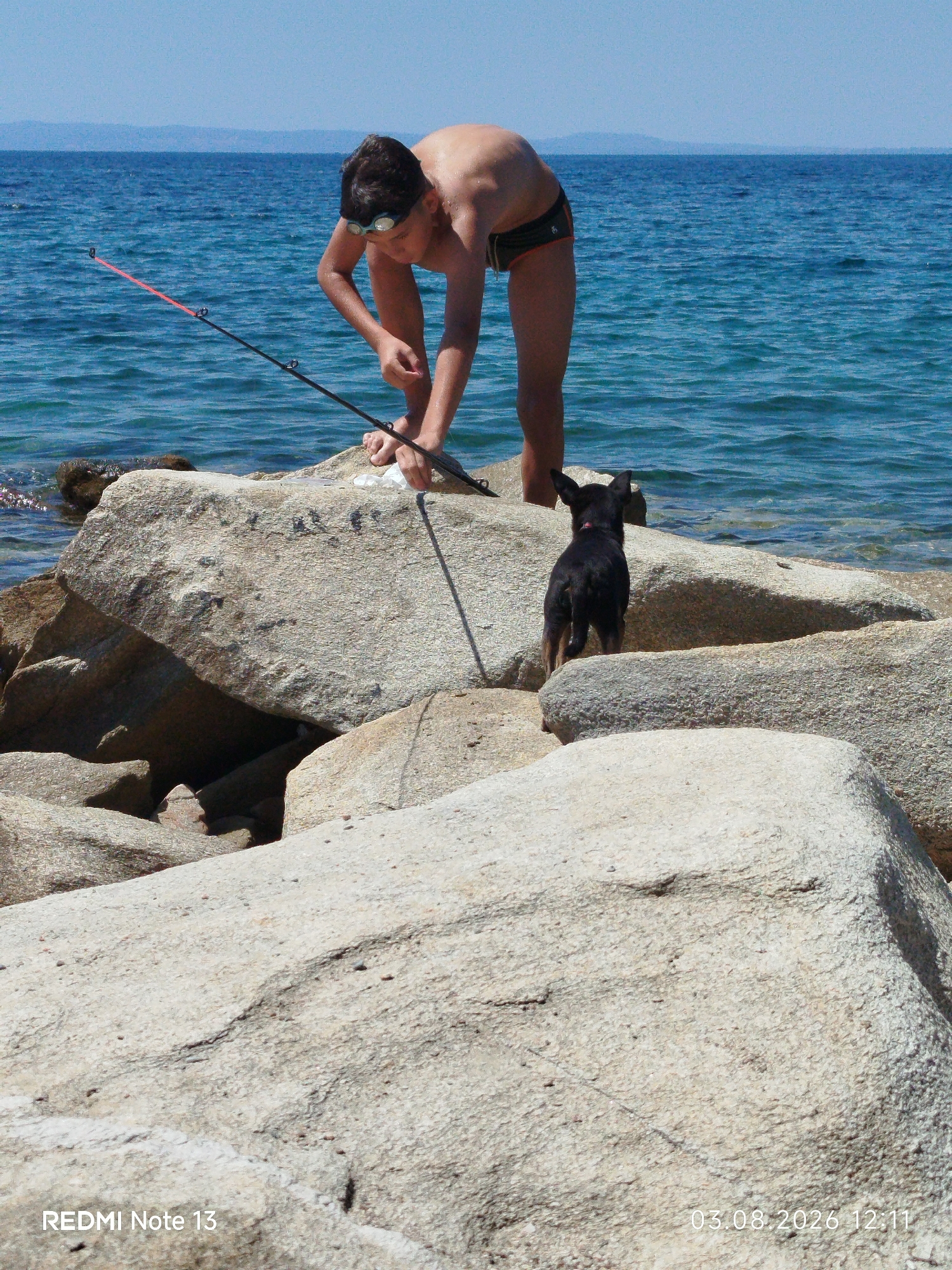 A boy with goggles on his forehead, wearing dark swim trunks, is fishing on a rocky shore. He is bent over, holding a fishing rod with a red tip, and appears to be attending to something near the water. A small black dog with tan paws is standing on the rocks behind him, looking out at the sea. The sea is blue and calm with a distant hazy coastline. The rocks are large and light-colored. The timestamp "03.08.2026 12:11" and "REDMI Note 13" are superimposed on the image. The setting is a sunny outdoor location, consistent with the coast of Sithonia Municipal Unit, Greece.