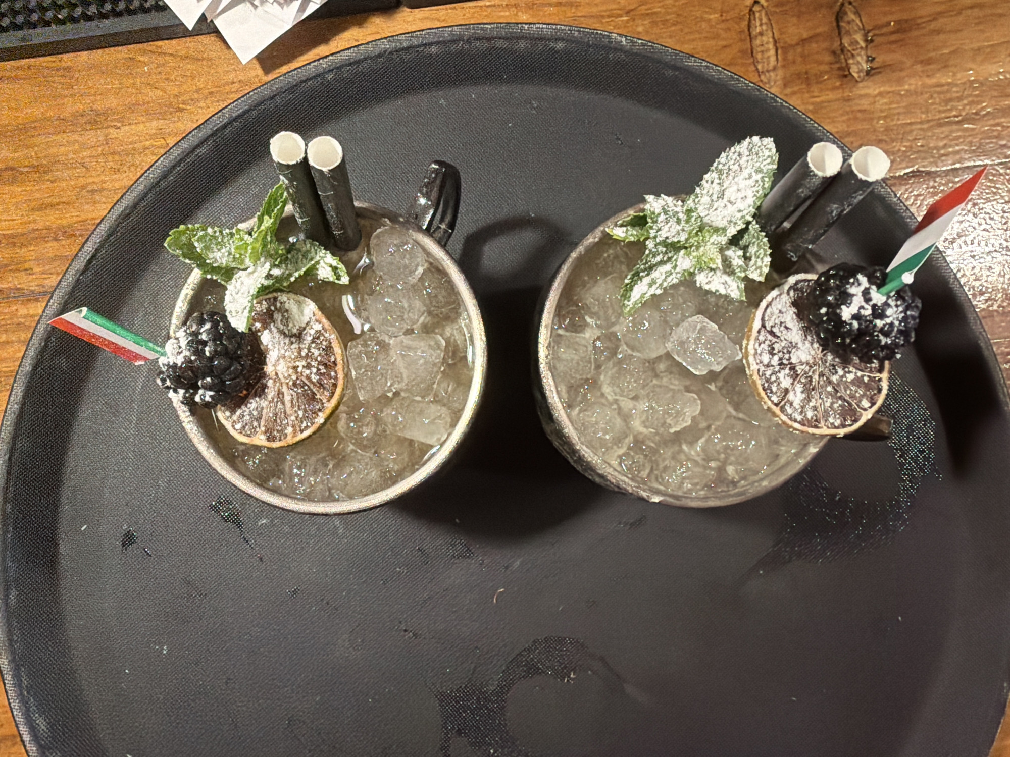The image shows two cocktails on a black tray. The tray is on a wooden surface. The cocktails are in silver cups and filled with ice. Each cocktail is garnished with a slice of dried orange, a blackberry, mint leaves, and two black straws. One cocktail also has a small flag with red, white, and green stripes. There is a small amount of powdered sugar on the garnishes. The overall scene is well-lit and appears to be taken from a top-down perspective. The location is not discernible from the image.