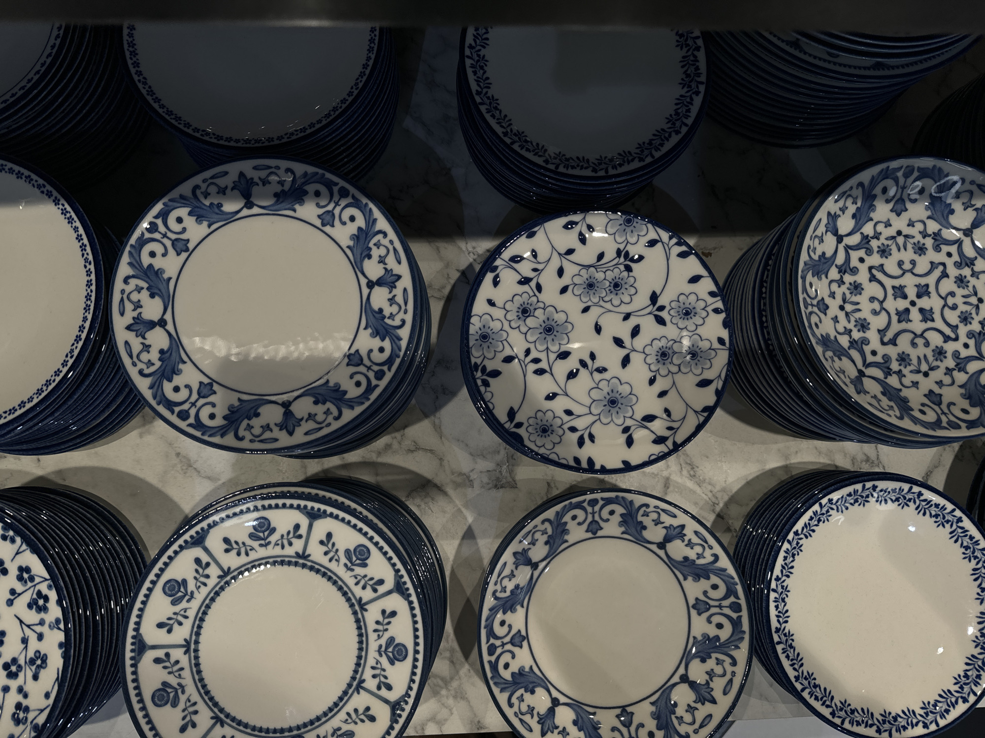 The image shows a close-up view of multiple stacks of white plates with blue floral patterns displayed on a shelf, likely in a store. The plates are arranged in neat stacks, suggesting an organized retail setting. The background appears to be a marble-like surface, indicating a possibly upscale environment. The location in the city of Yudino, Russia cannot be determined from the image.