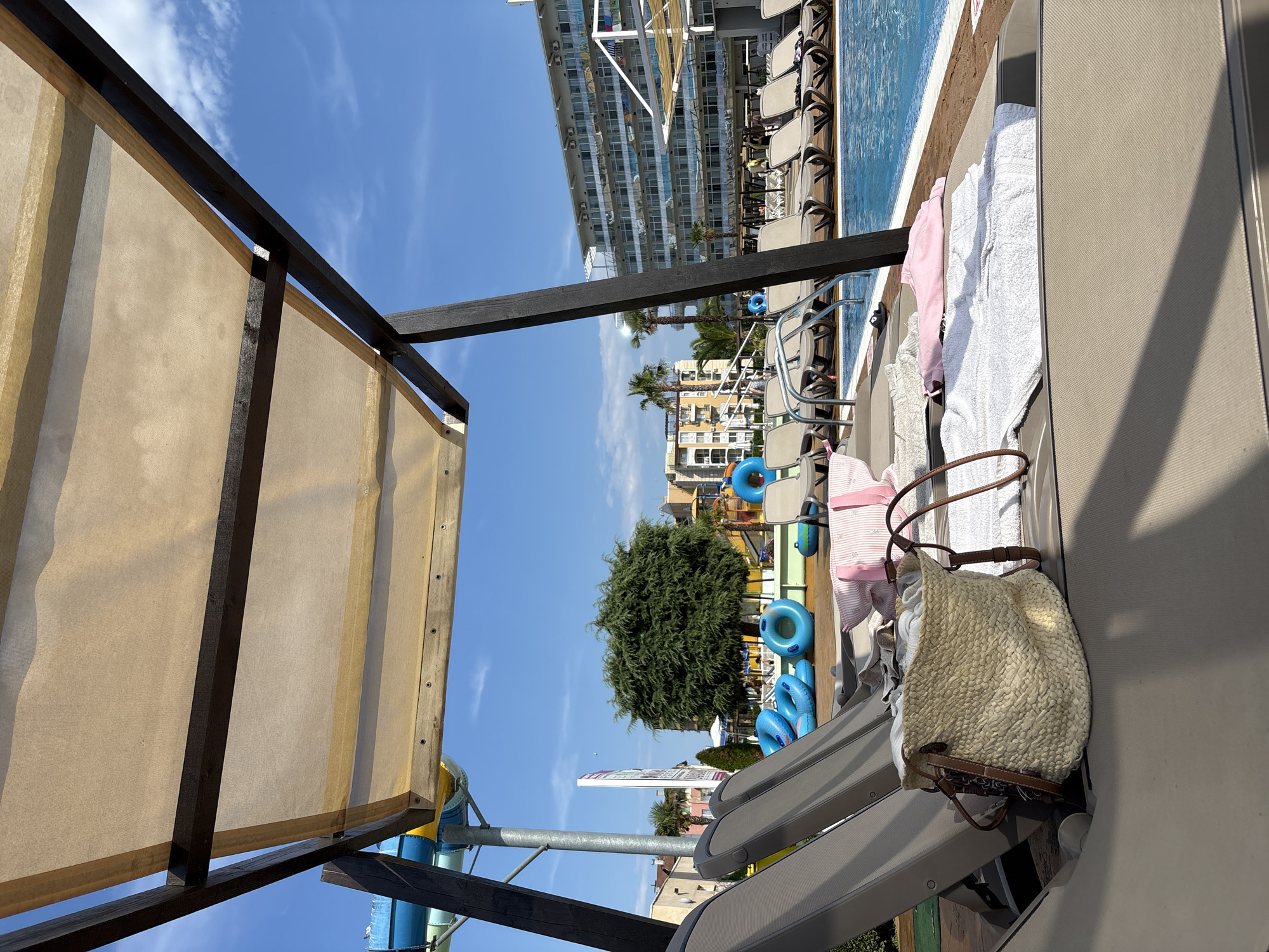 The image captures a sunny day at an outdoor swimming pool complex, likely in Nesebar, Bulgaria. In the foreground, several lounge chairs are set up under a tan canopy supported by dark wooden beams. A woven straw bag and folded towels, one pink and one white, are placed on one of the chairs, suggesting a relaxed atmosphere. In the middle ground, the bright blue water of the swimming pool is visible, with more lounge chairs lining its edge. Beyond the pool, a large, modern hotel building with numerous balconies stands under a clear blue sky. Various water slides and inflatable rings, some blue, are scattered around the pool area, indicating a family-friendly resort. Lush green trees and other buildings are visible in the background, contributing to the resort-like ambiance. The scene appears to be a typical vacation setting, with an emphasis on leisure and outdoor recreation.