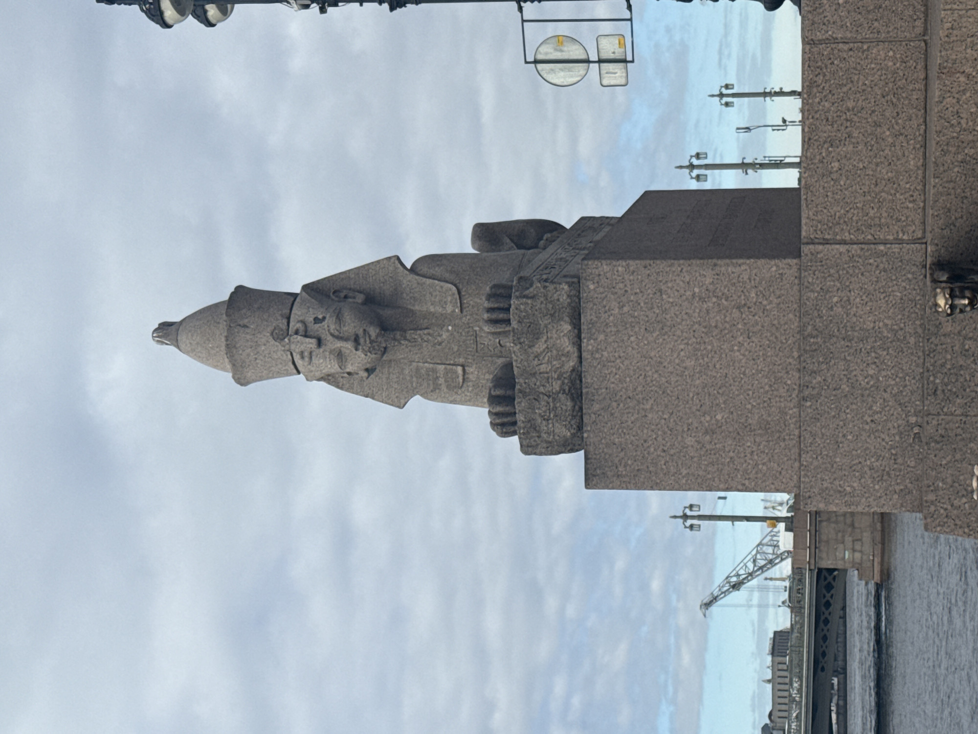 The image depicts a griffon statue, located on the Bridge of Griffins, a pedestrian bridge in Saint Petersburg, Russia. The griffon is sculpted in gray stone, featuring a human face and the body of a mythical creature. The statue is affixed to a stone base. The background includes a cloudy sky, a portion of the bridge structure, street lamps, and a building further down the river.