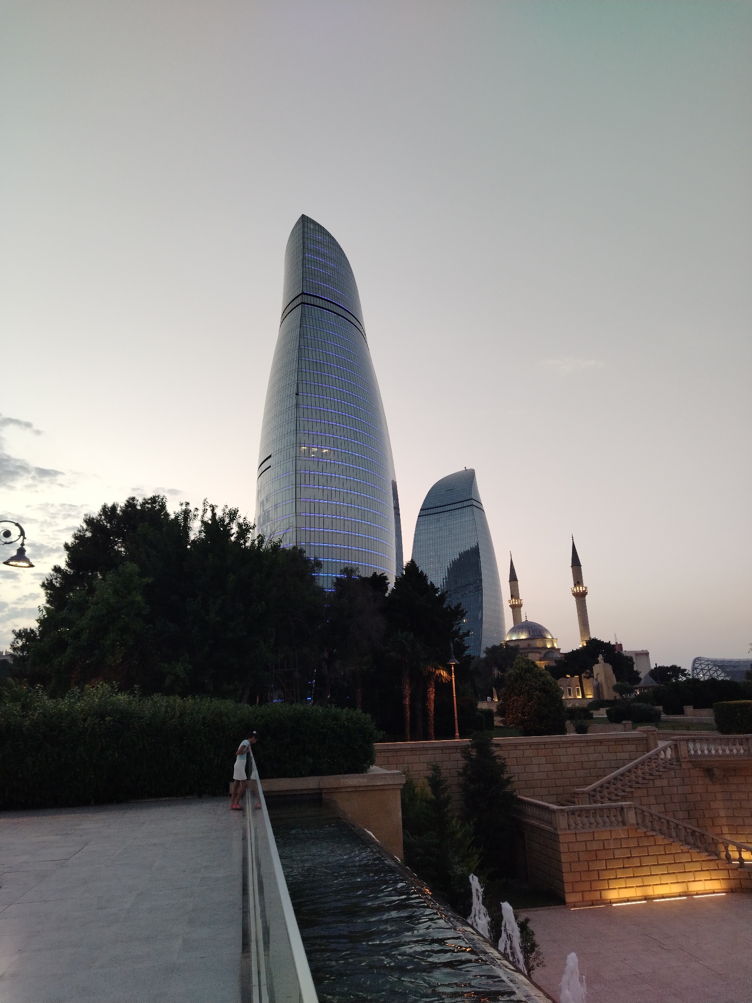 The image captures a scene in Sabail Raion, Azerbaijan, featuring modern skyscrapers and traditional architecture at dusk. In the background, two prominent flame-shaped skyscrapers, illuminated with blue lights, dominate the skyline. To their right, a mosque with a dome and two minarets is visible, also lit up. In the foreground, a person is standing on the edge of a walkway overlooking a water feature with small fountains. The walkway is bordered by a low, manicured hedge. Beyond the hedge, a stone staircase with decorative railings ascends, leading to a plaza area. The sky is a soft grey, transitioning to a lighter hue near the horizon, suggesting either early morning or late evening. Lush trees and architectural elements contribute to the layered urban landscape.