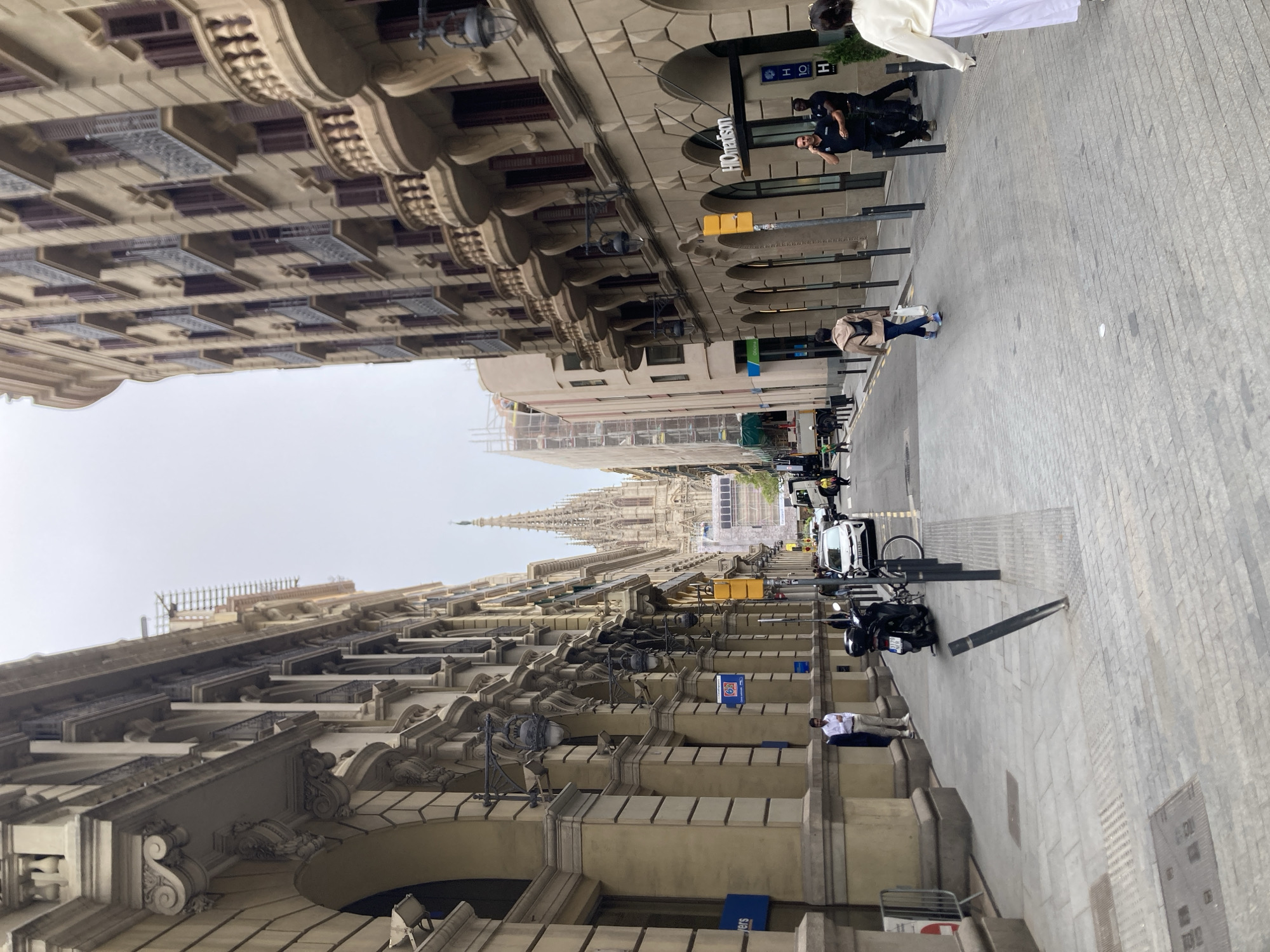 The image shows a street in Barcelona, Spain. The street is lined with ornate buildings featuring detailed architectural facades. At the end of the street, the spires of a cathedral can be seen. There are people walking and standing on the pavement. Several parked motorcycles and cars are visible along the street. A hotel, identified by a sign, is also present.