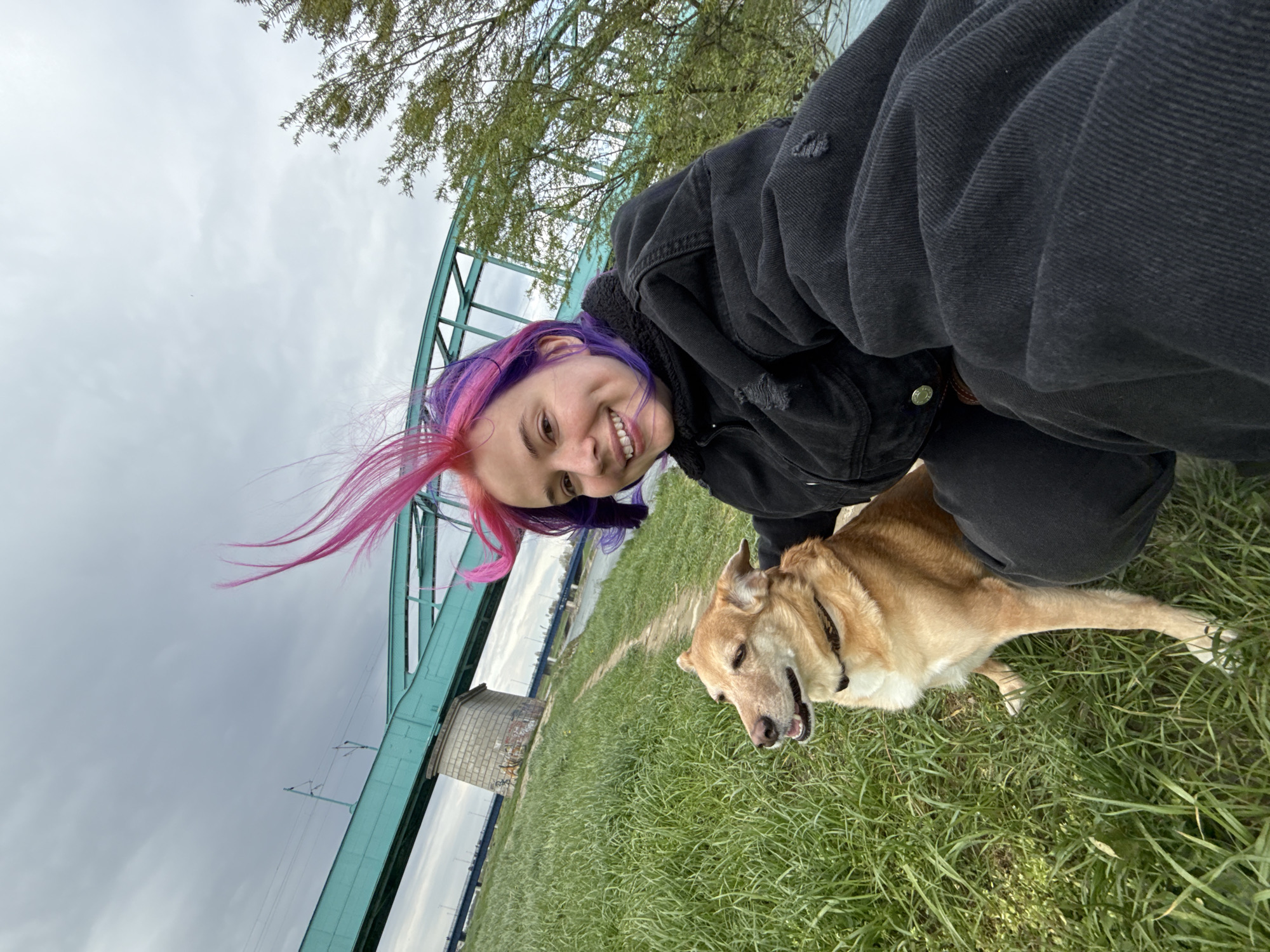 A tilted, close-up shot shows a young woman with brightly colored hair (pink and purple) crouching next to a light brown dog in a grassy area. The woman is smiling at the camera. She is wearing a dark jacket with a light-colored collar. The dog appears to be panting slightly. Behind them, a light blue bridge is visible, along with a section of a river and a cloudy sky. A tree branch with leaves is also present. The location is in the city of Zagreb, Croatia.