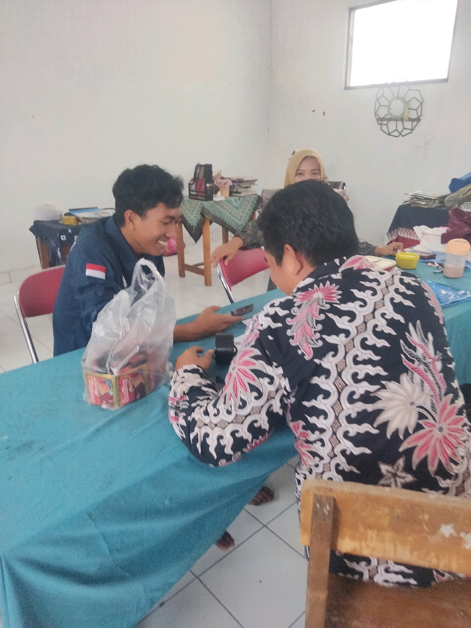 In a room that appears to be a classroom or meeting space in Suka Sari, Indonesia, three individuals are seated around a teal-colored table covered with a tablecloth. On the table, to the left, is a clear plastic bag containing what appears to be packaged snacks, next to a box with red and gold accents. A man in a dark blue uniform shirt with a white and red stripe on the left sleeve is laughing and looking at a mobile phone, which he is holding in his right hand. He is sitting opposite another individual. Another person, wearing a black batik shirt with a white and pink floral pattern, is seated and looking down at their hands. A woman wearing a beige headscarf is visible in the background, between the two individuals at the table. The room has white walls and a window on the right side, which is brightly lit. In the background, there are stacks of papers, a black wire rack, and a decorative circular object mounted on the wall.