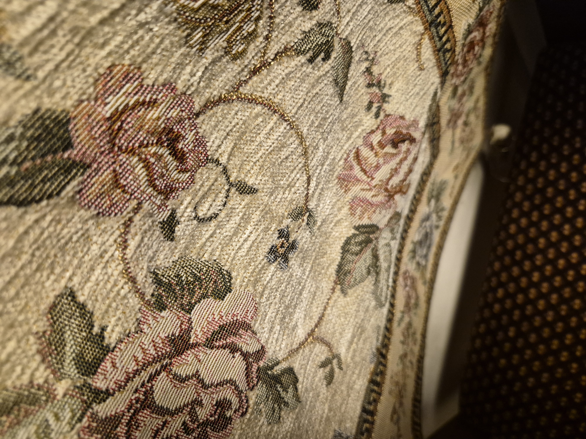 This is a close-up, low-angle shot of a patterned fabric, possibly a tablecloth or upholstery, with a floral motif. The fabric has a textured weave with threads of beige, cream, gold, pink, and dark green. The floral designs include roses and leaves. A dark, patterned surface, likely carpet or a similar material, is visible in the lower portion of the image, out of focus. A white surface is partially visible between the patterned fabric and the dark lower surface, suggesting a piece of furniture. The image does not contain any information that would allow for its localization in Vanadzor, Armenia, or any other specific geographical location.